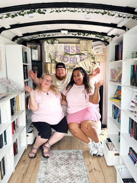 Three people sitting in a bookstore aisle, smiling and waving, surrounded by bookshelves, with a sign reading 'SISS FORD' behind them.