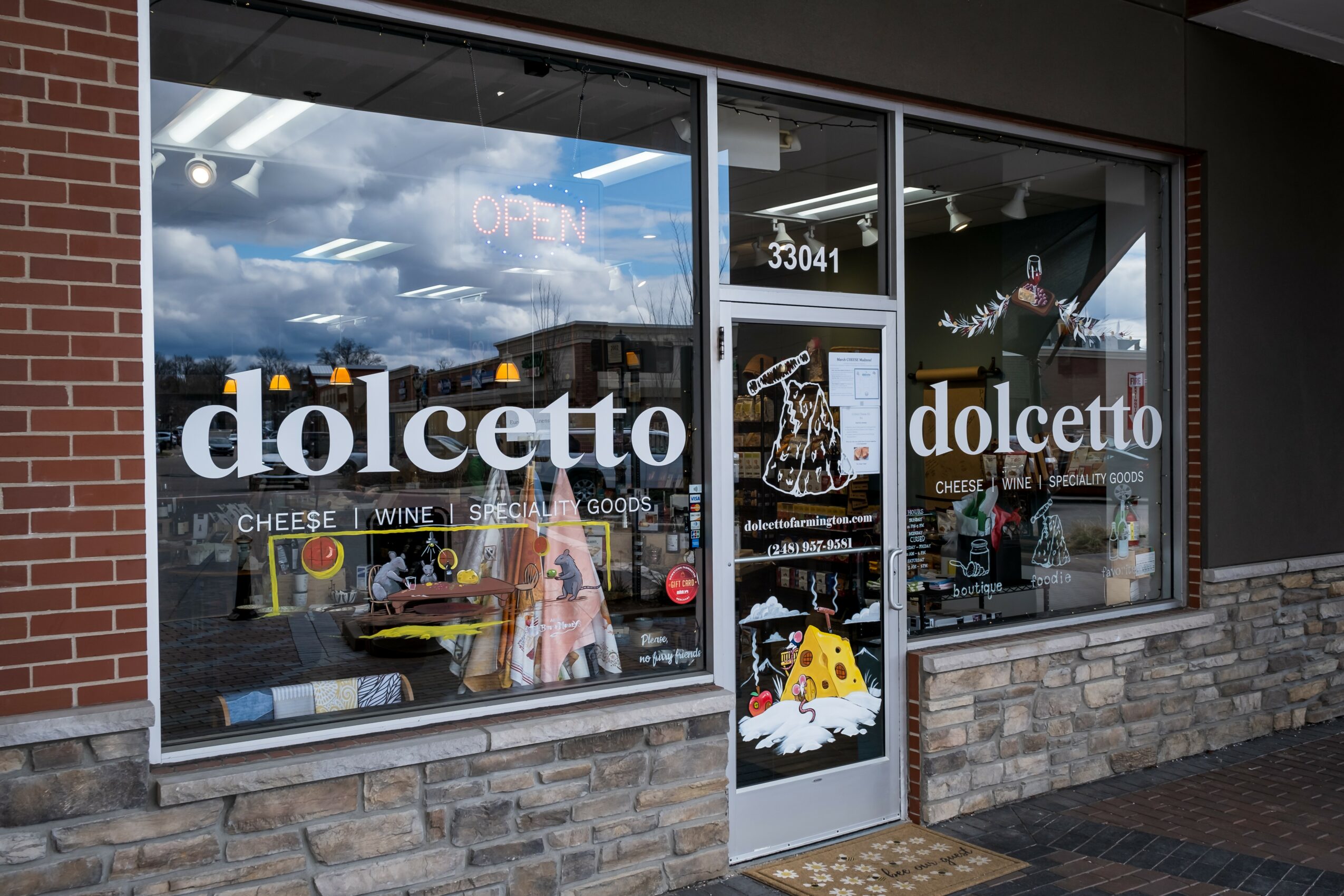 Storefront with large glass windows and door, displaying the name 'dolcetto' and a cheese and wine sign, brick and stone exterior.