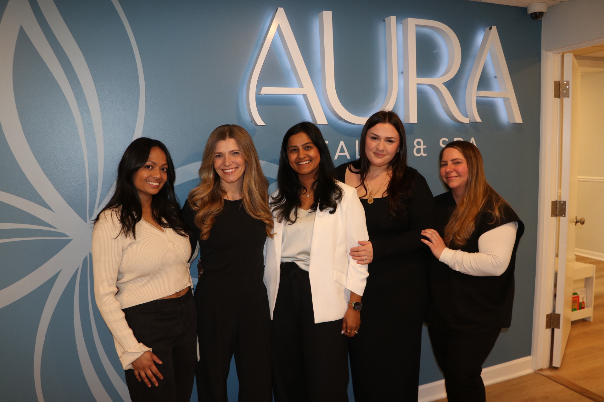 Five women standing together in front of a blue wall with illuminated text 'AURA'. They are smiling and dressed in black and white clothing.