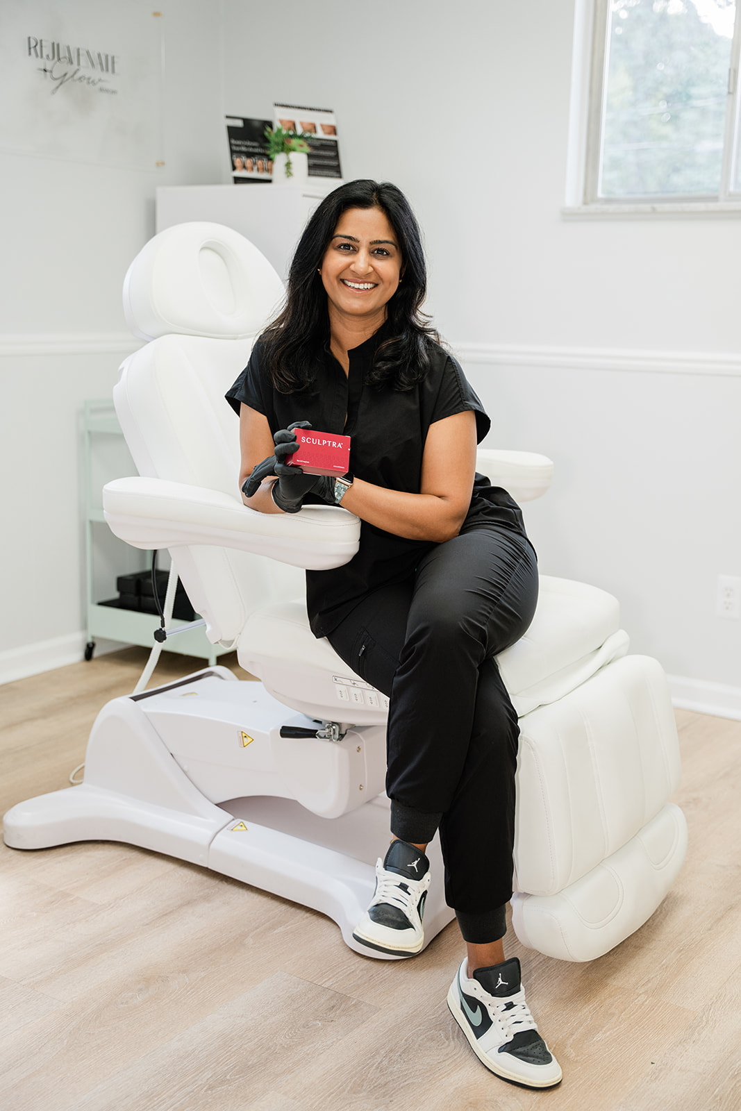 Woman sitting on a white medical chair, smiling, holding a small red device, in a bright room with a window.