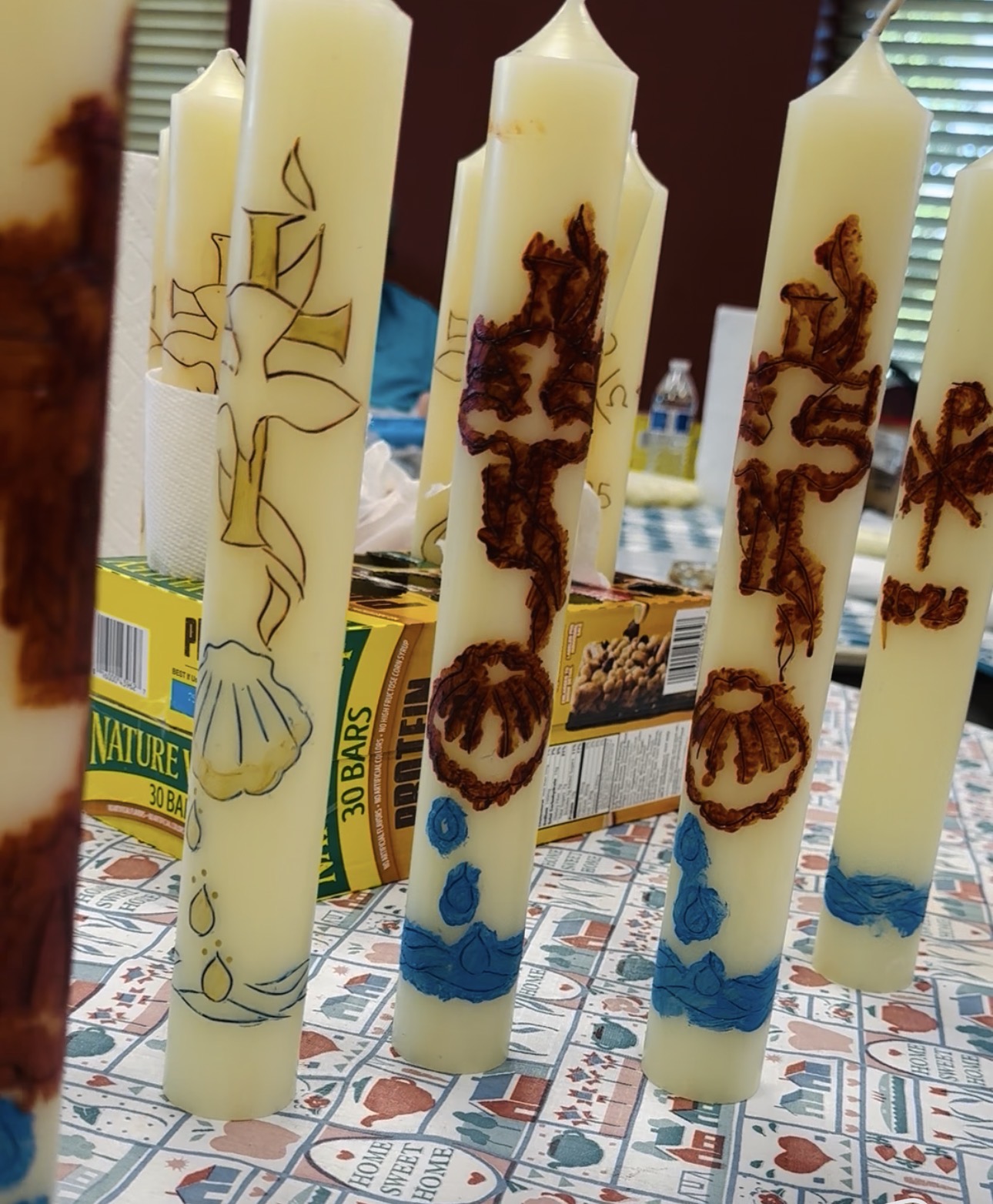 Tall white candles with brown and blue decorative patterns, placed on a patterned surface with boxes in the background.