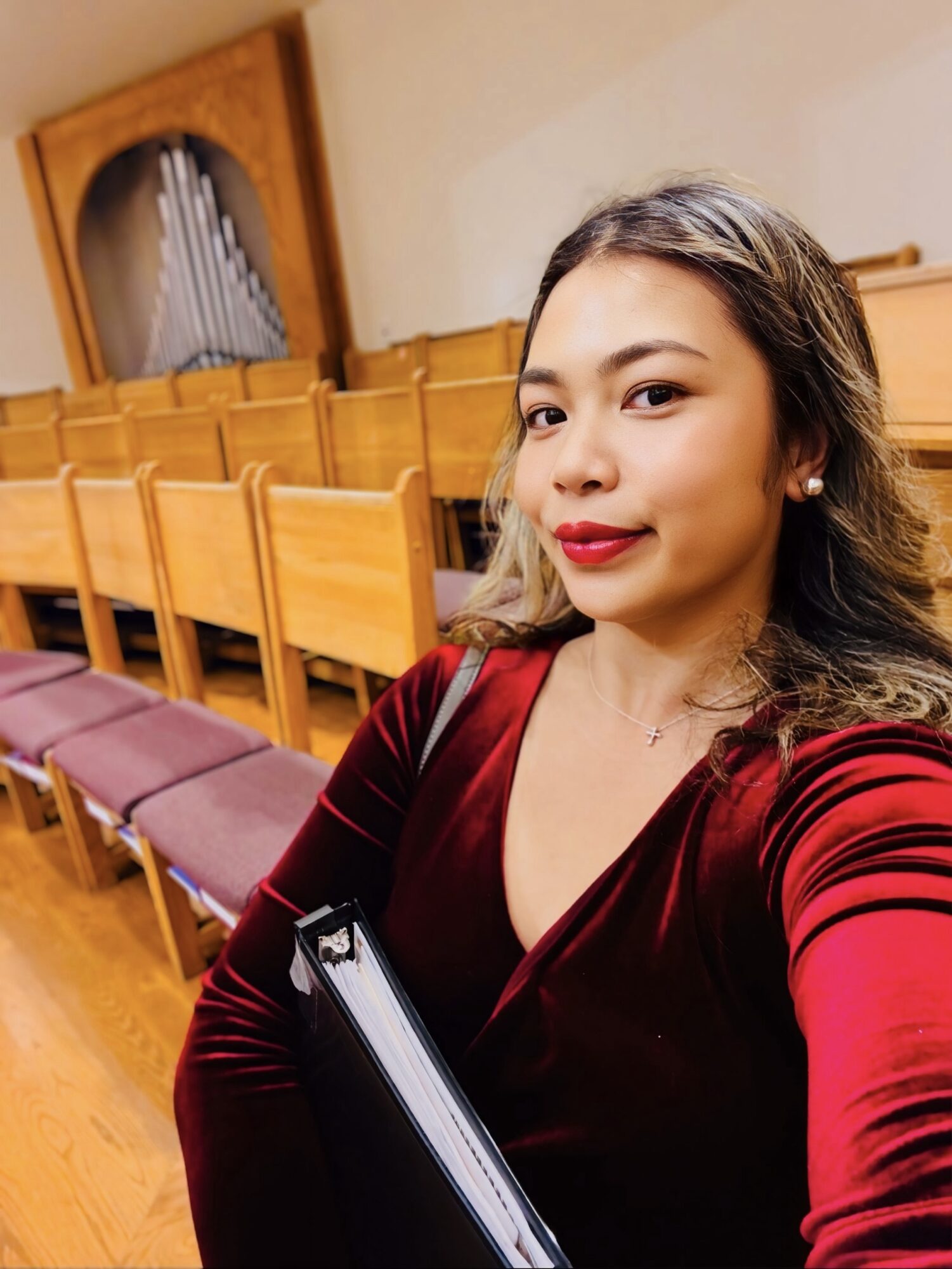 Young woman with wavy hair and red lipstick taking a selfie in a room with wooden chairs and a pipe organ in the background.