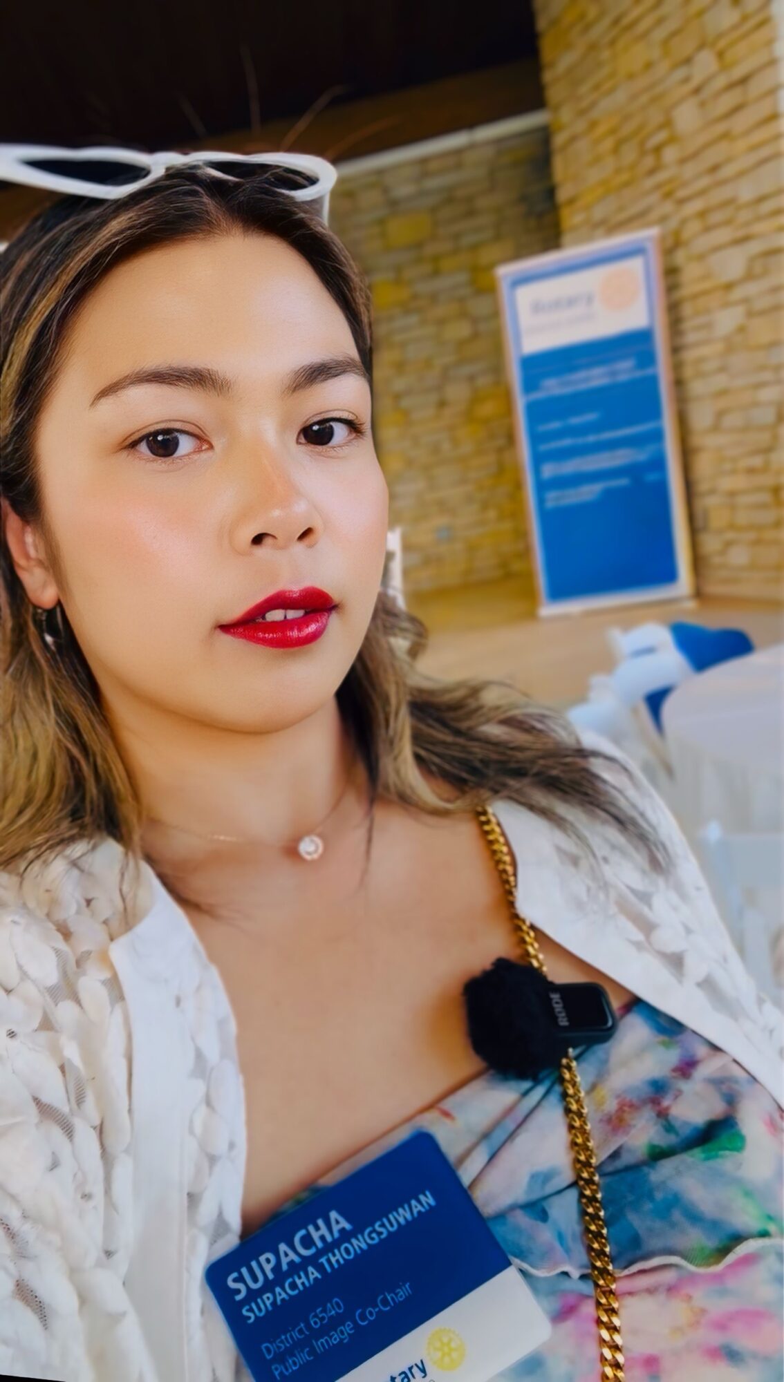 Young woman with sunglasses on head, red lipstick, and a colorful top, standing indoors near a blue informational sign.