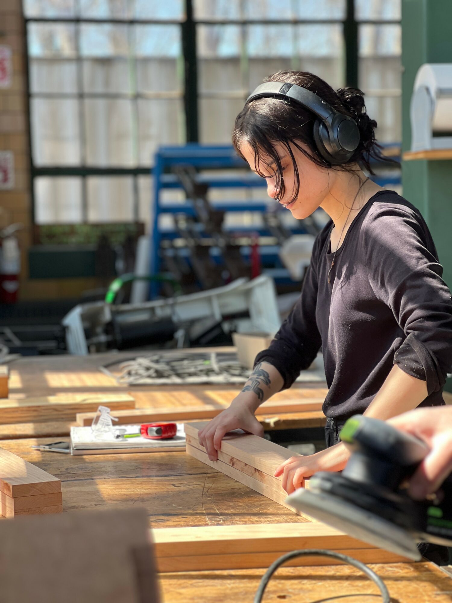 Woman wearing headphones working on a woodworking project in a workshop, using a measuring tool on a wooden board.