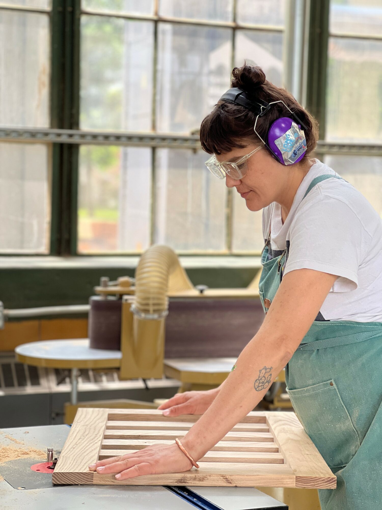 Woman wearing headphones and glasses working on a wooden board in a bright room with large windows.