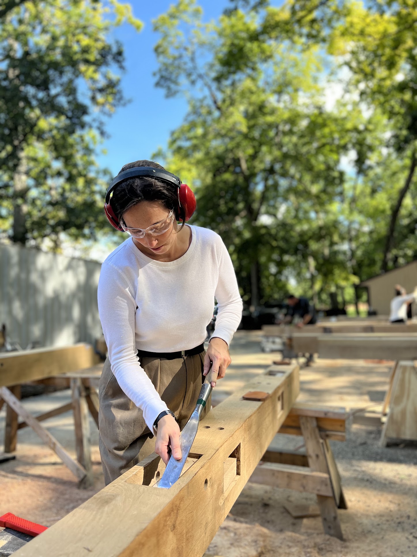 Person wearing safety glasses and headphones, using a saw on a wooden beam outdoors, surrounded by trees and benches.