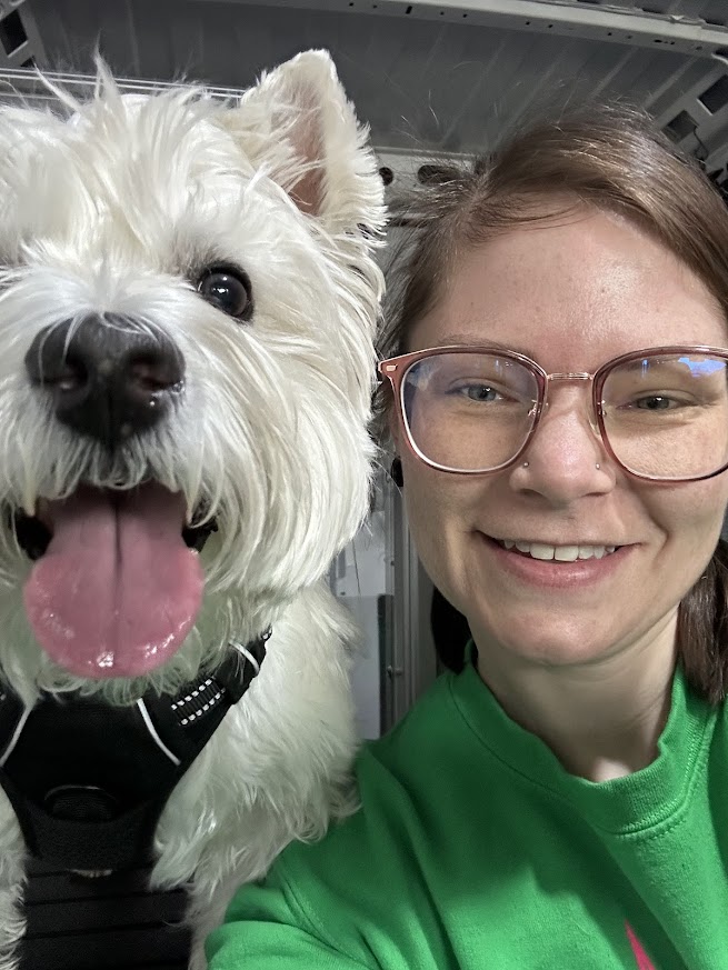 Person with glasses smiling next to a white dog with open mouth and tongue out, both close-up.