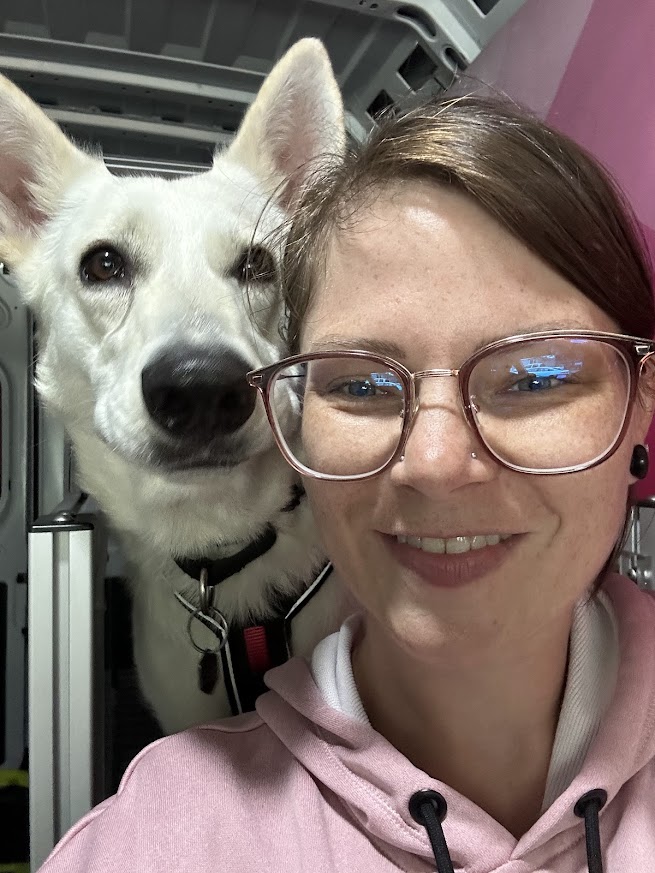Woman with glasses smiling next to a white dog with pointy ears, in an indoor setting.