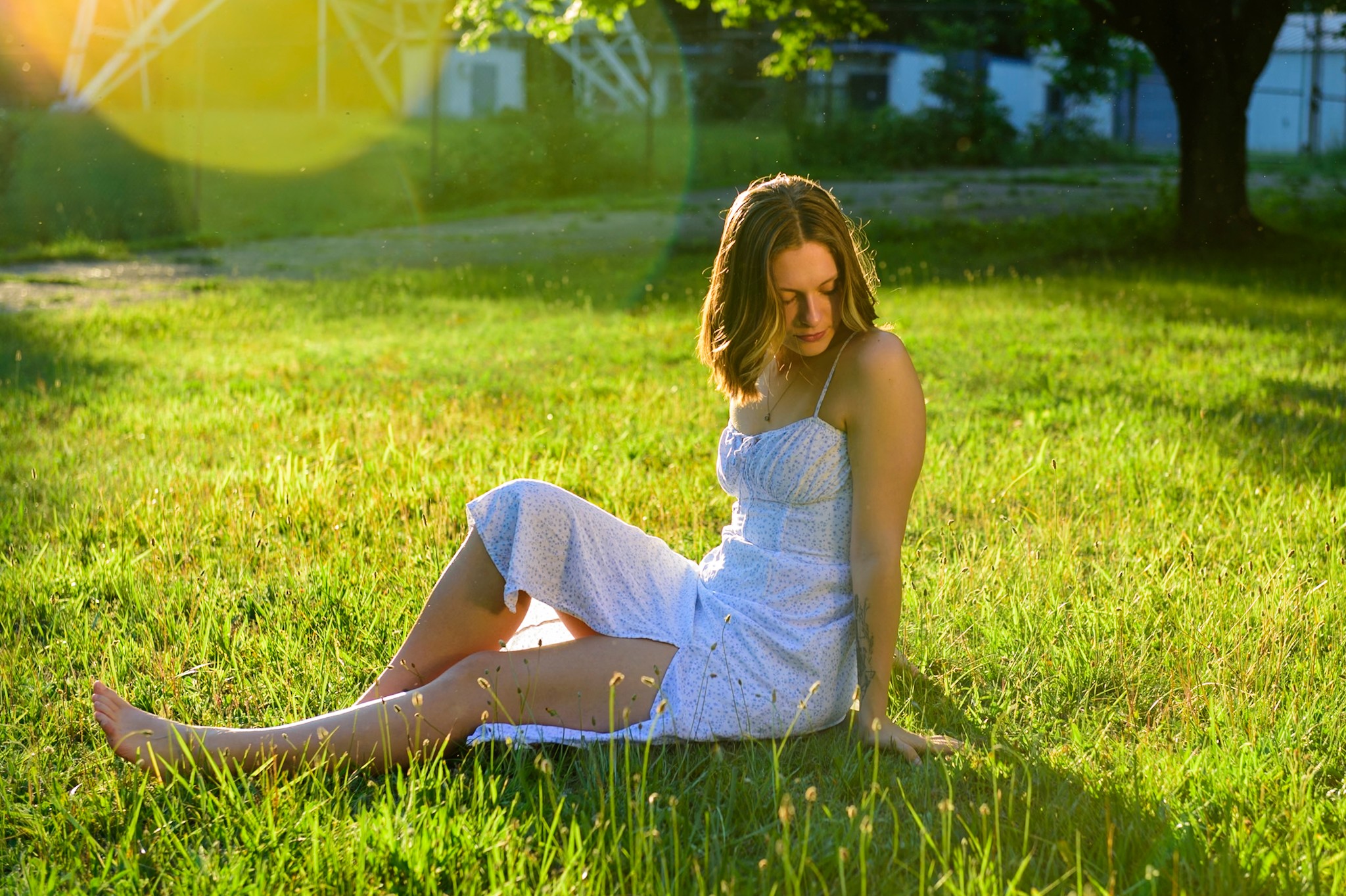 Woman in a white dress sitting on grass in sunlight, with trees and a park background.