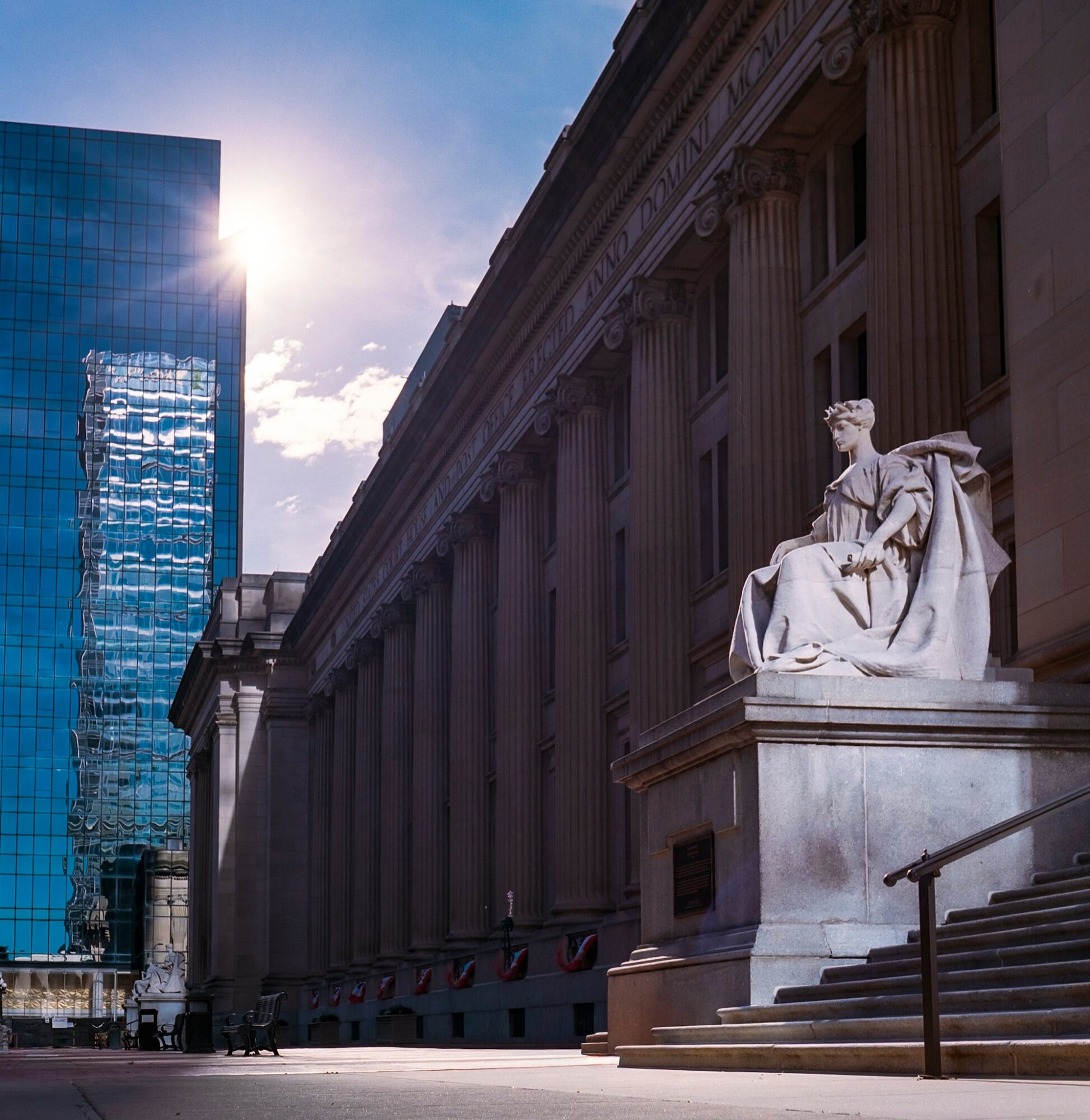 A classical building with columns and a seated statue in front, sunlight shining from the left, modern glass building nearby.