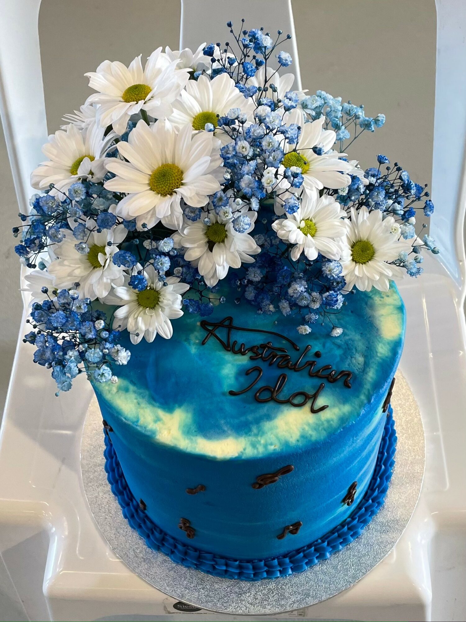 Blue cake decorated with white daisies and blue baby's breath flowers, with handwritten text and small black musical notes, on a silver base.