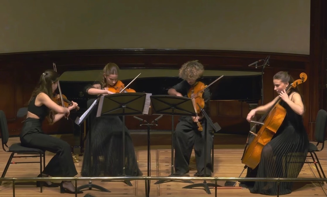 Four young musicians playing string instruments on stage, seated in a semi-circle, with music stands in front of them.