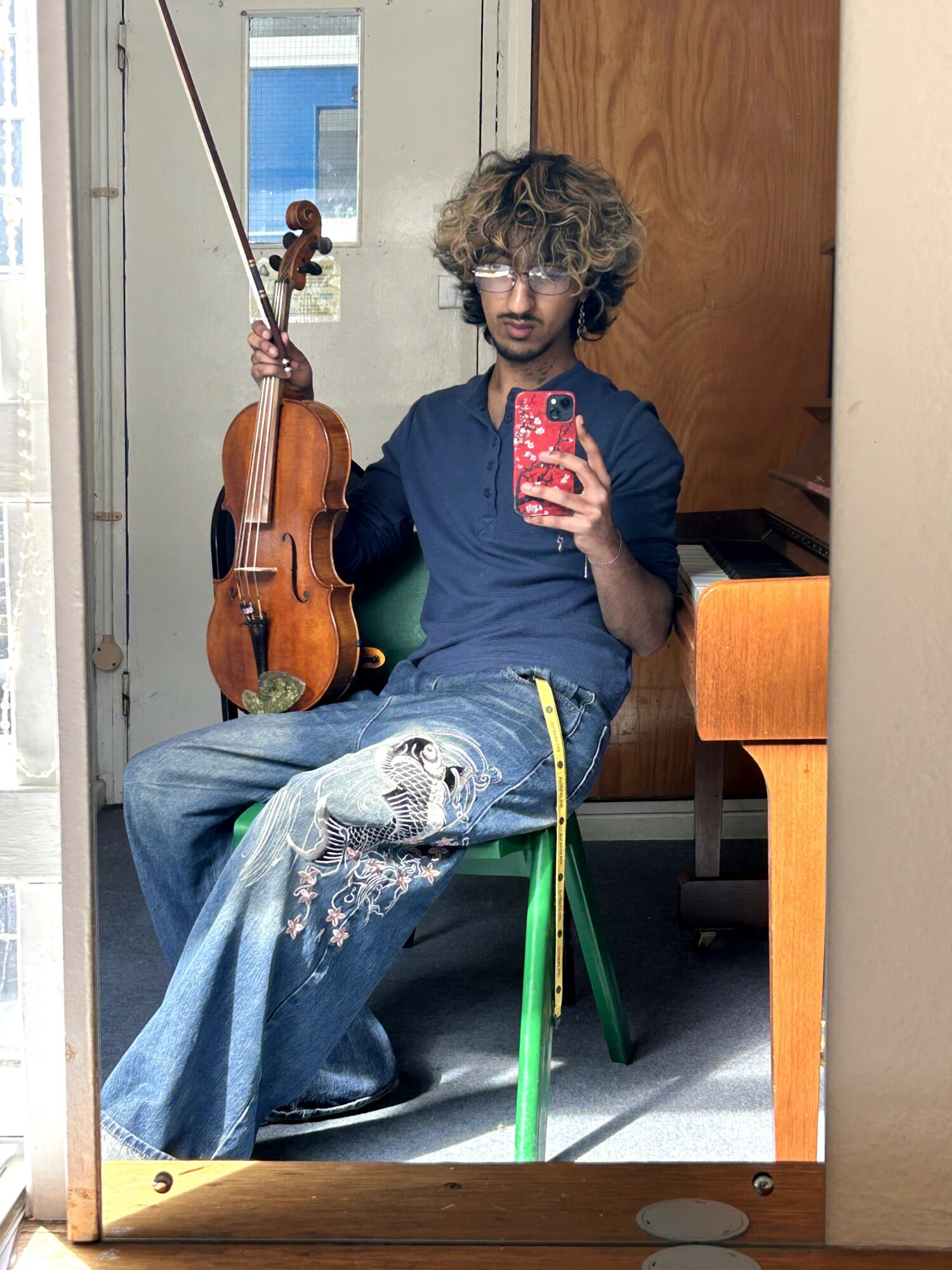 Person with curly hair taking a mirror selfie, holding a violin, sitting on a chair, wearing a dark shirt and jeans with paint splatters.