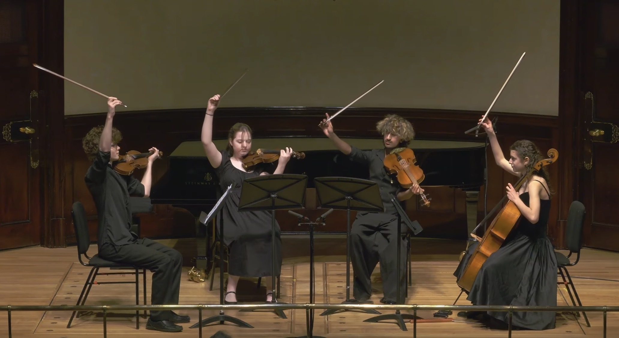 Five musicians perform on stage, playing violin and cello, with a large screen behind them.