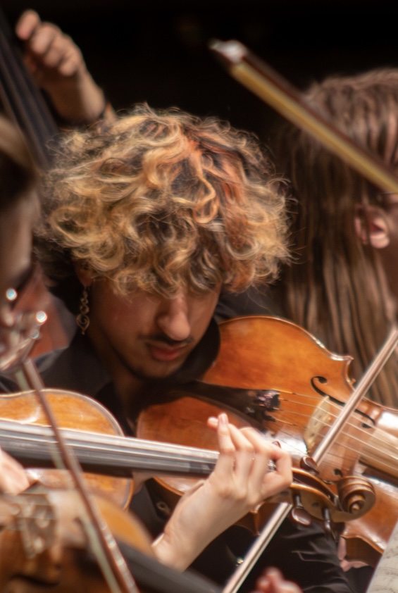 Person with curly hair playing violin in an orchestra, surrounded by other musicians, in a dark setting.