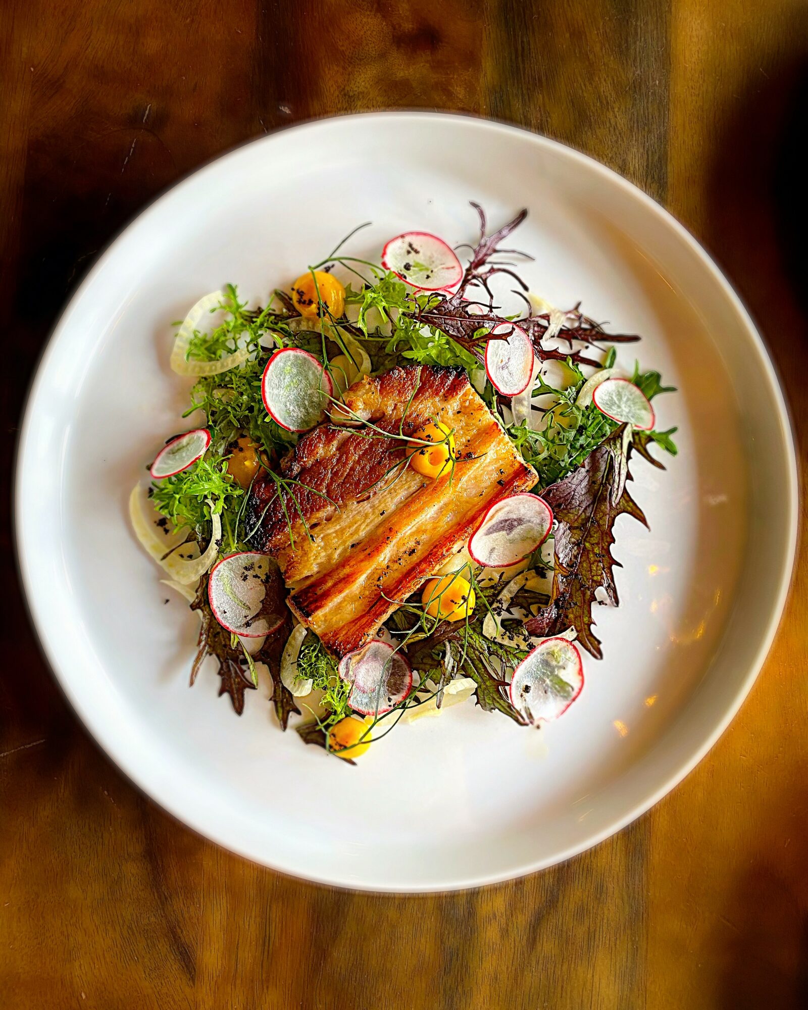 Salad with greens, radish slices, and herbs on a white plate, placed on a wooden surface.