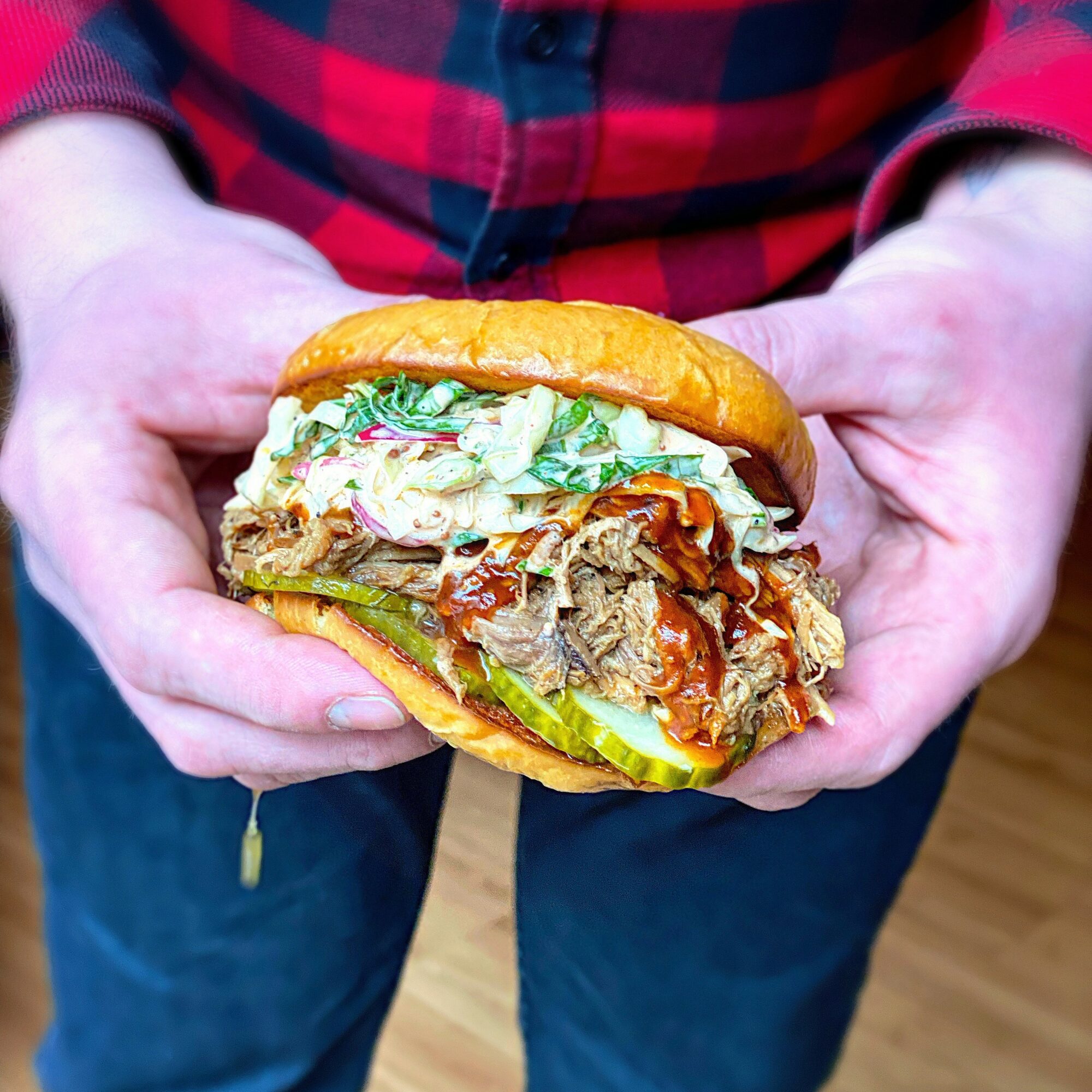 Person holding a large sandwich with meat, lettuce, and sauce in a bun, with hands visible and a red plaid shirt.