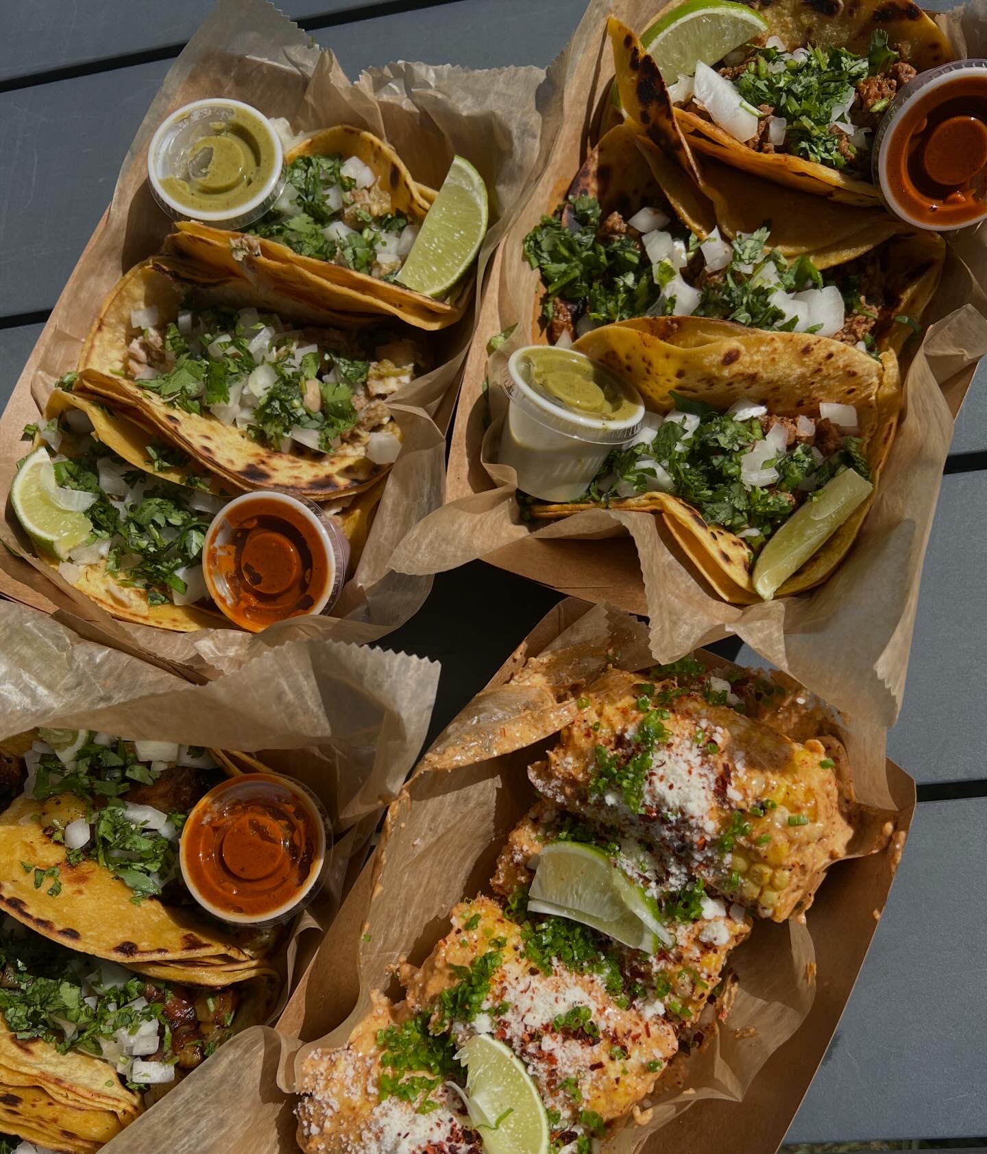 Four tacos with lime wedges, sauces, and toppings on brown paper, on a dark surface.