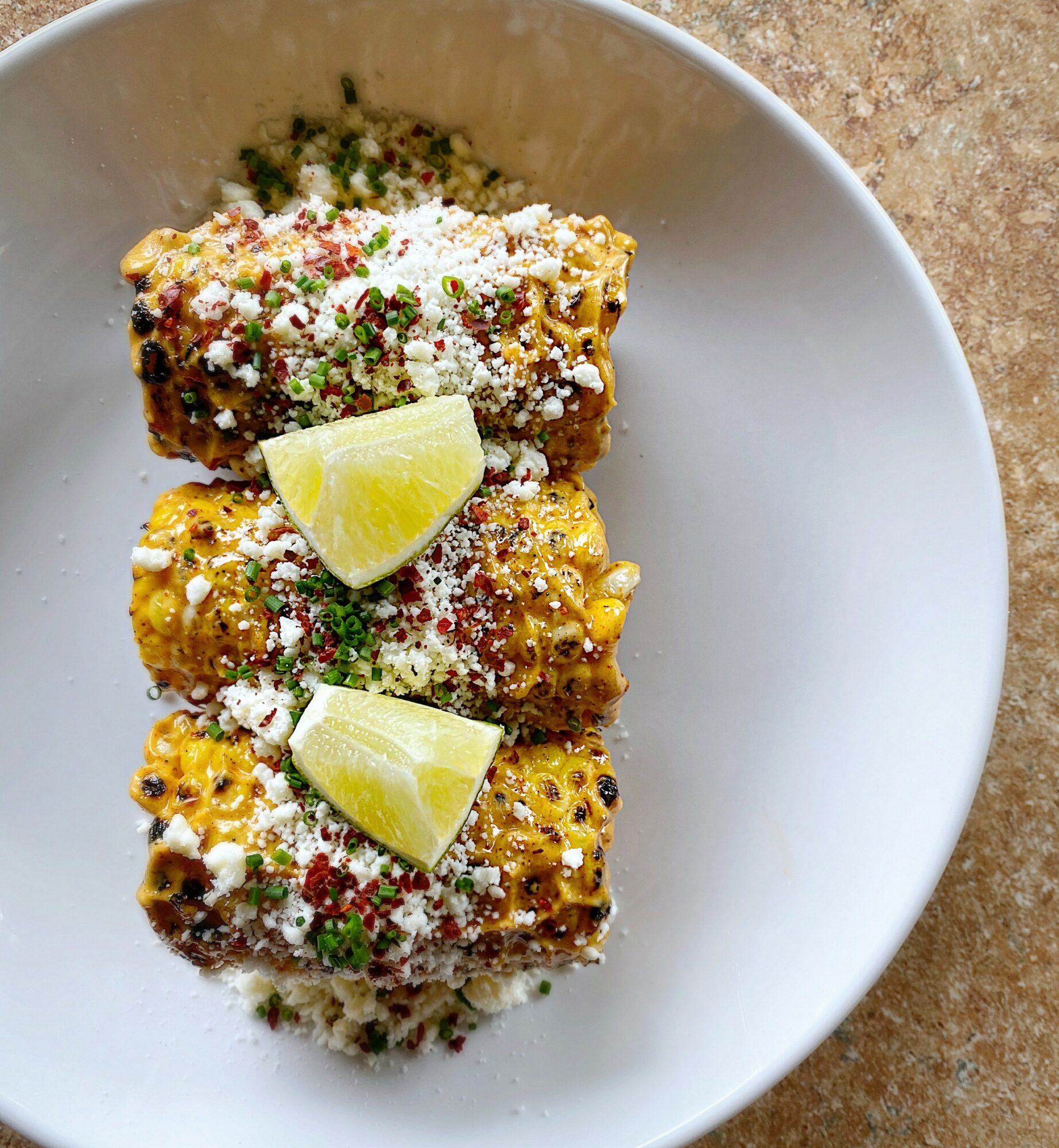 Three pieces of Mexican street corn topped with cheese, chili powder, and lime wedges on a white plate.