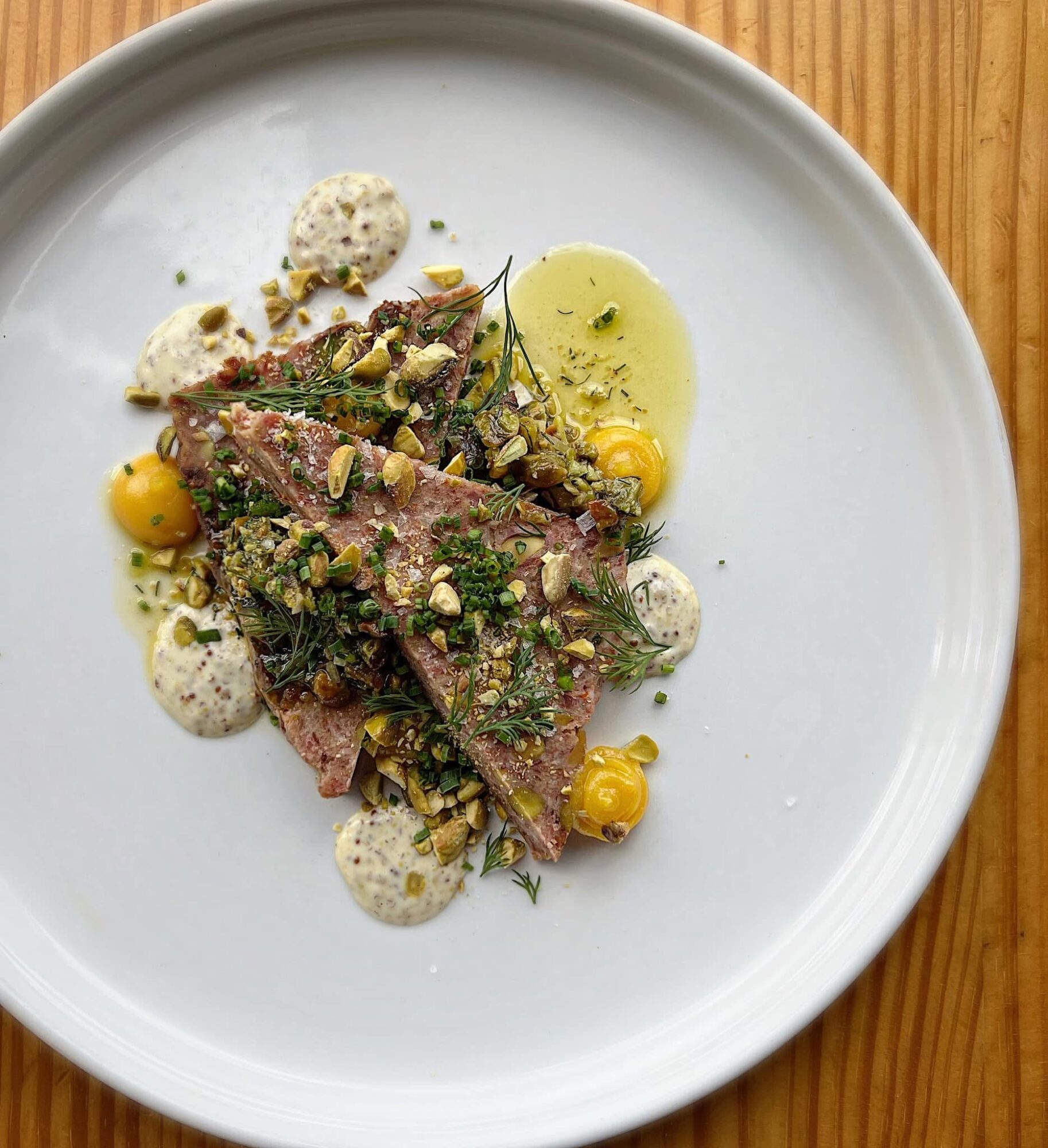Plate with sliced meat, yellow dots, green herbs, and white sauce on a white dish, wooden table background.