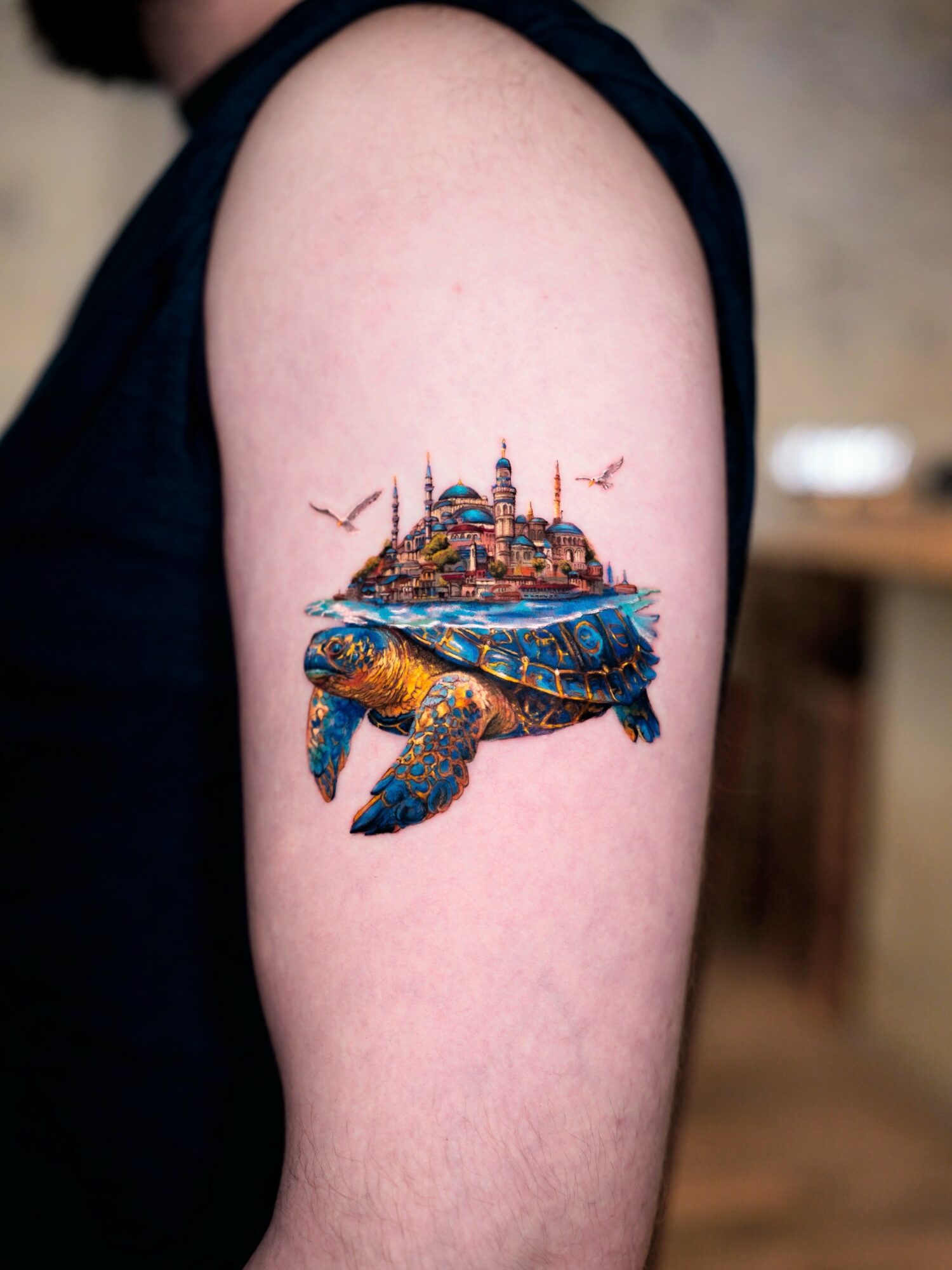 Colorful turtle tattoo with a cityscape on its back, on person's upper arm.