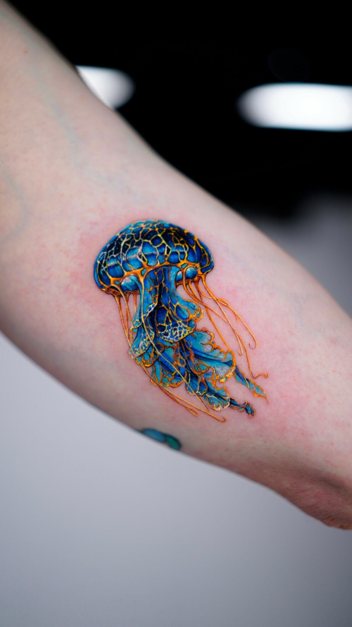 Colorful jellyfish tattoo on person's arm with blue, orange, and yellow details, and tentacles hanging down.