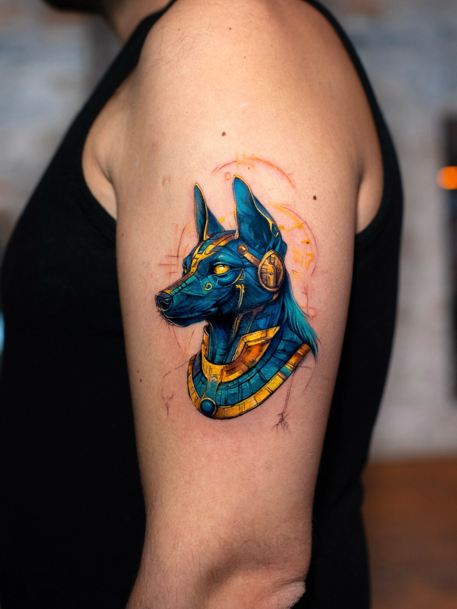 Tattoo of a blue Egyptian-style cat with gold jewelry on upper arm, detailed and colorful.