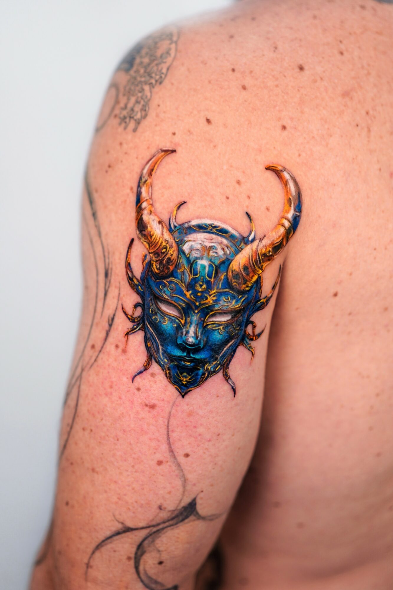 Tattoo of a blue and gold horned animal head on person's upper arm, with detailed features and shading.