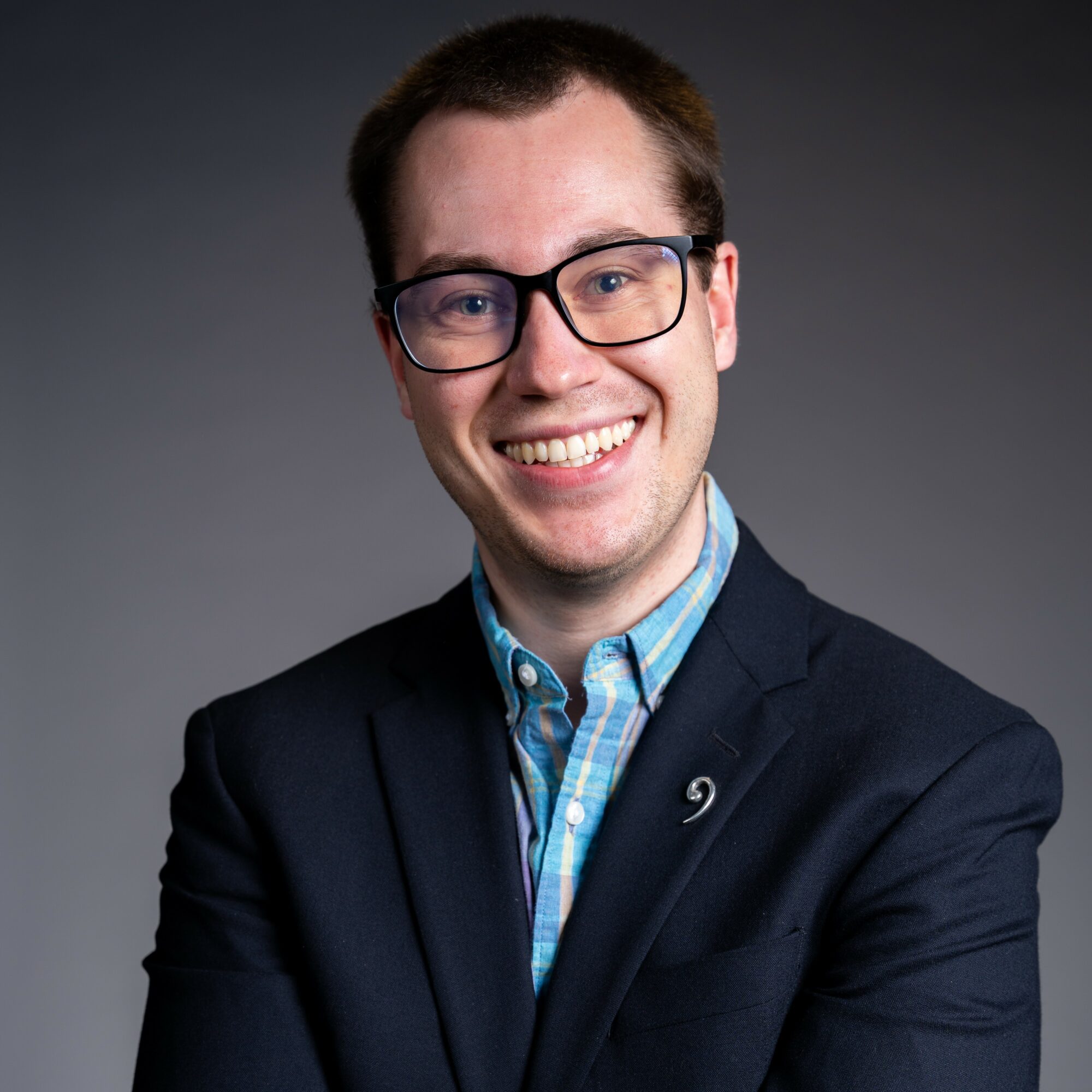 Smiling man with glasses, wearing a dark blazer and blue checkered shirt, against a gray background.