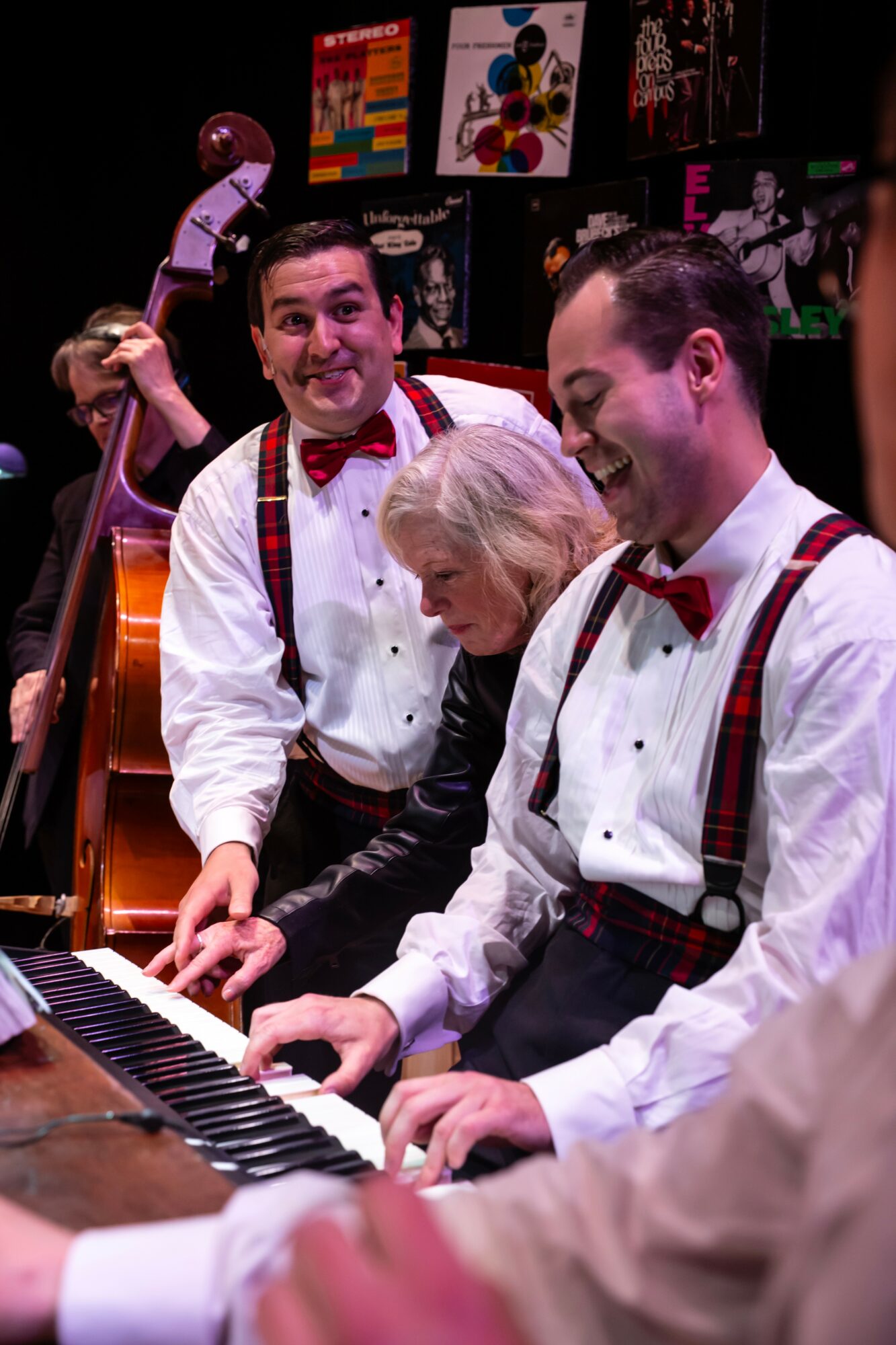 Three people playing piano, two men in white shirts with red bow ties and suspenders, woman with white hair, colorful posters in background.