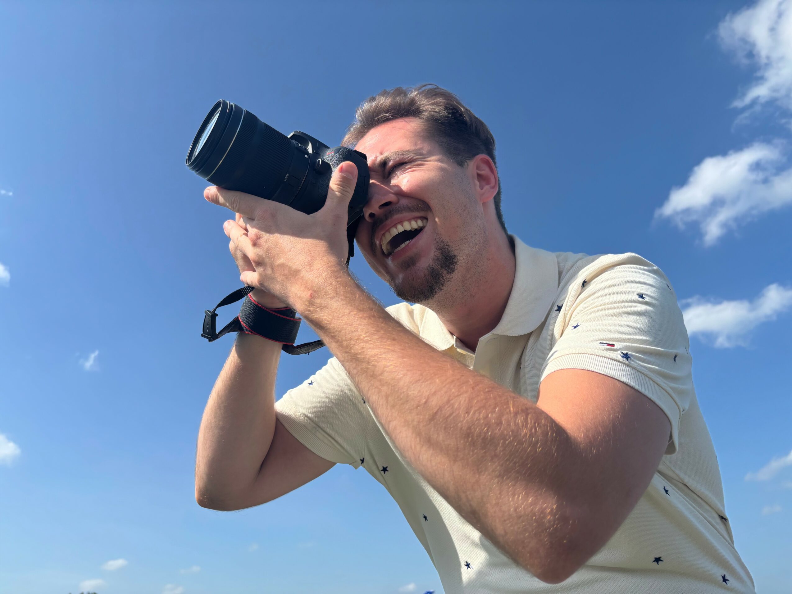 Man with short hair and beard taking a photo with a camera outdoors, smiling, against a blue sky with clouds.