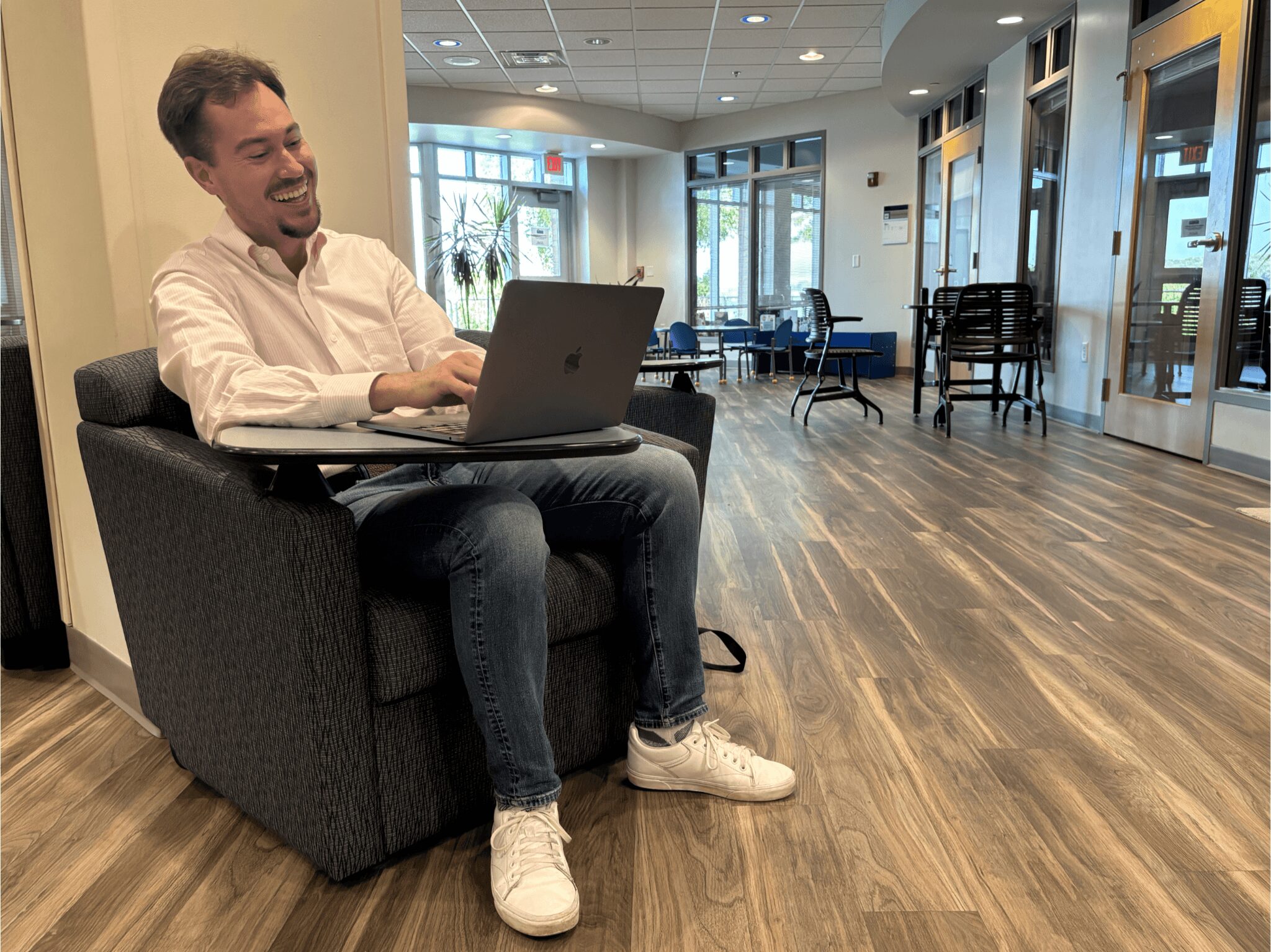 Person sitting on a black armchair using a laptop, smiling, in a modern lobby with wooden floors and glass doors.
