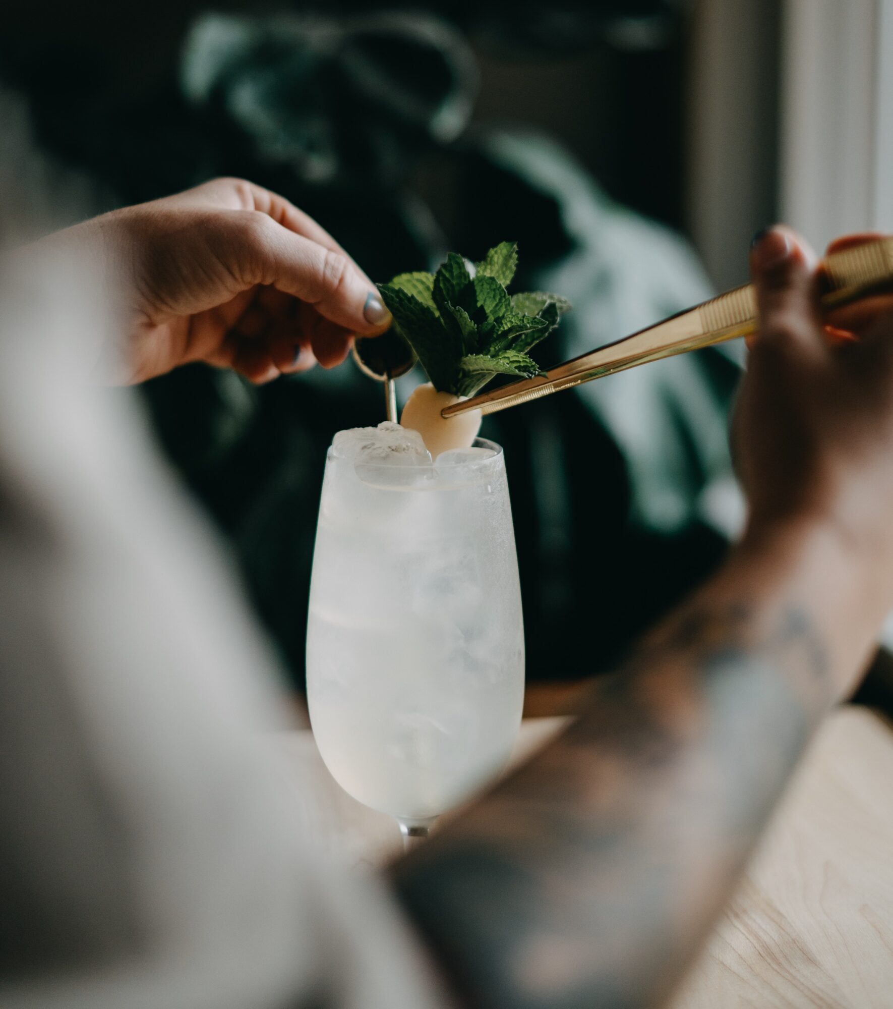 Person adds mint leaves to a tall glass with a straw, drink with ice, near a window, tattooed arm visible.