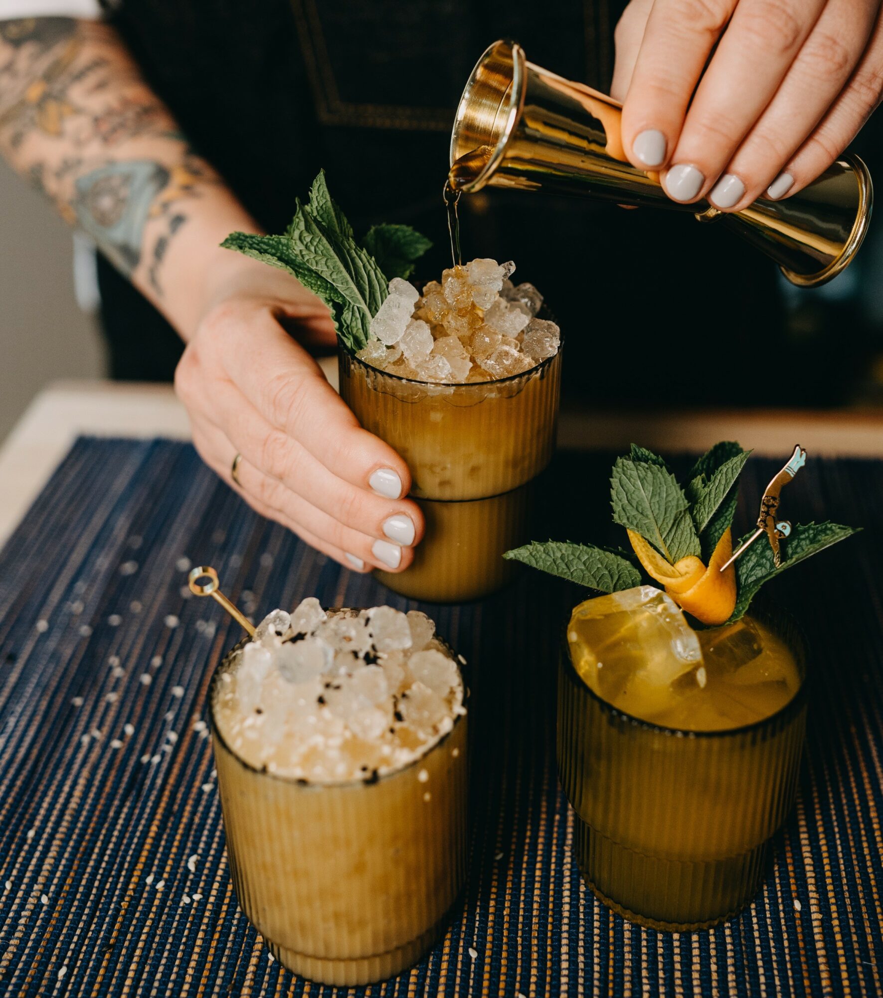 Person pouring liquid into a glass with crushed ice, mint leaves, and a garnish, on a dark table.