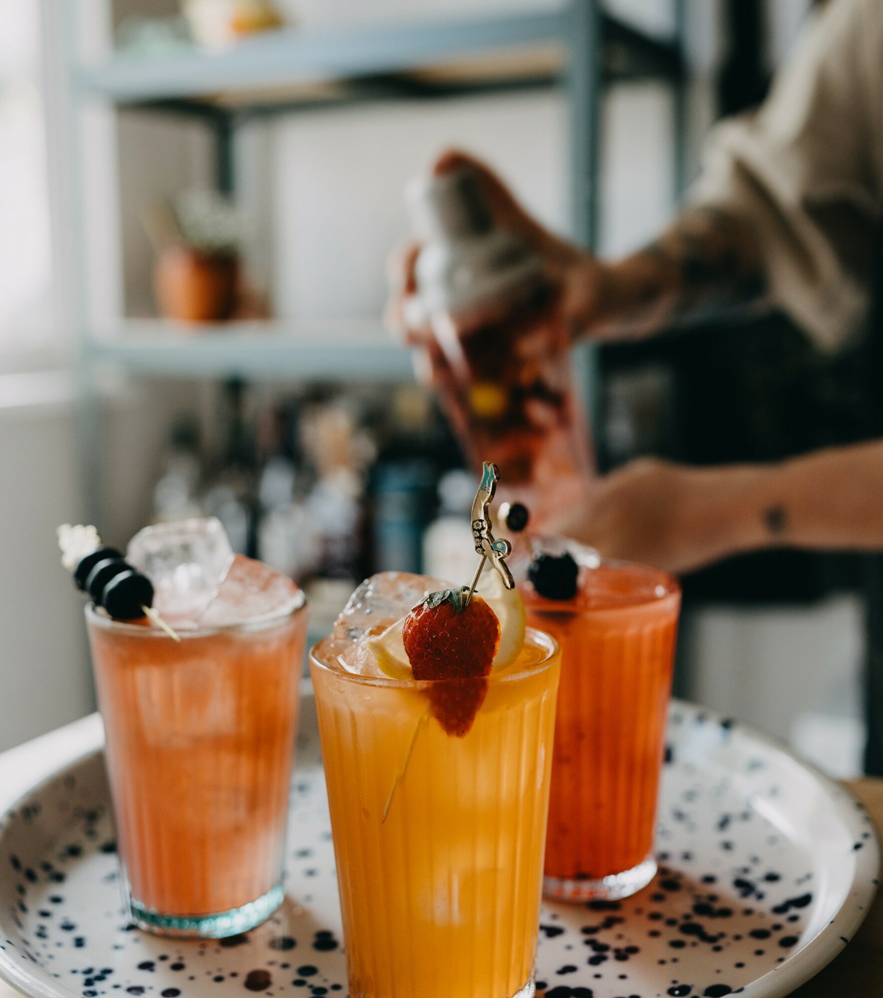 Three colorful drinks with ice and garnishes on a patterned tray, with a person pouring a drink in the background.