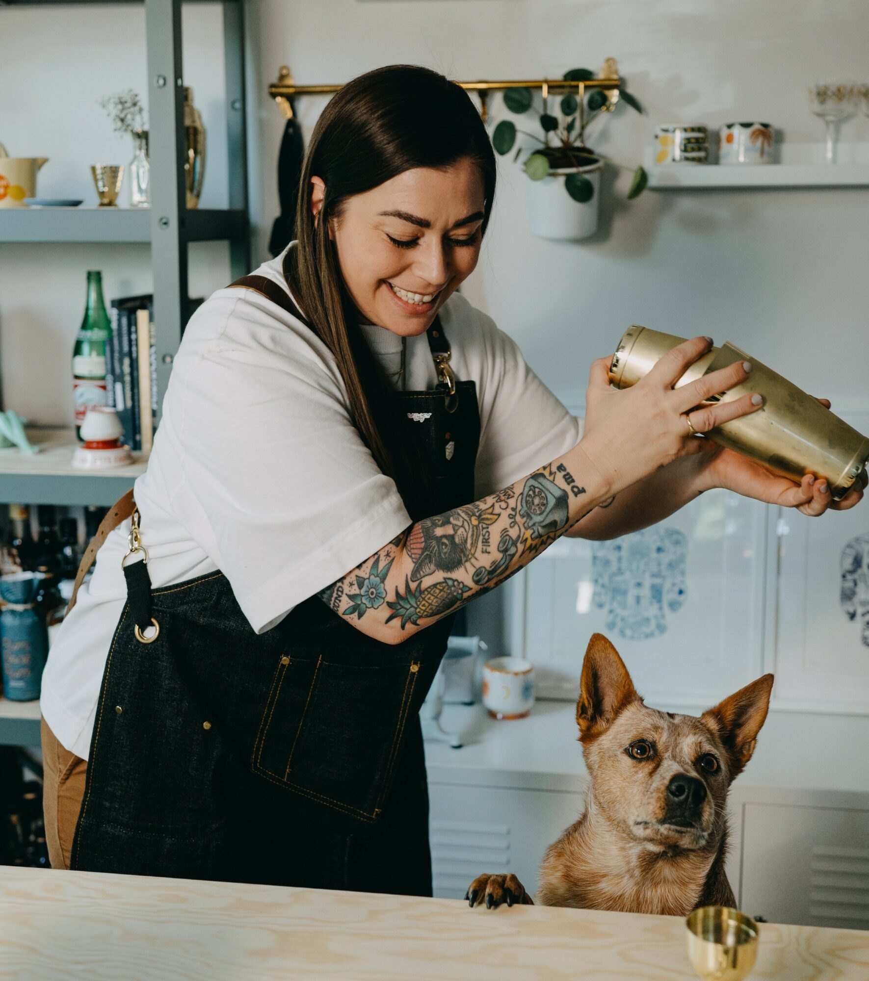 Woman smiling and pouring a drink into a glass, dog sitting on table nearby, kitchen background, bright lighting.