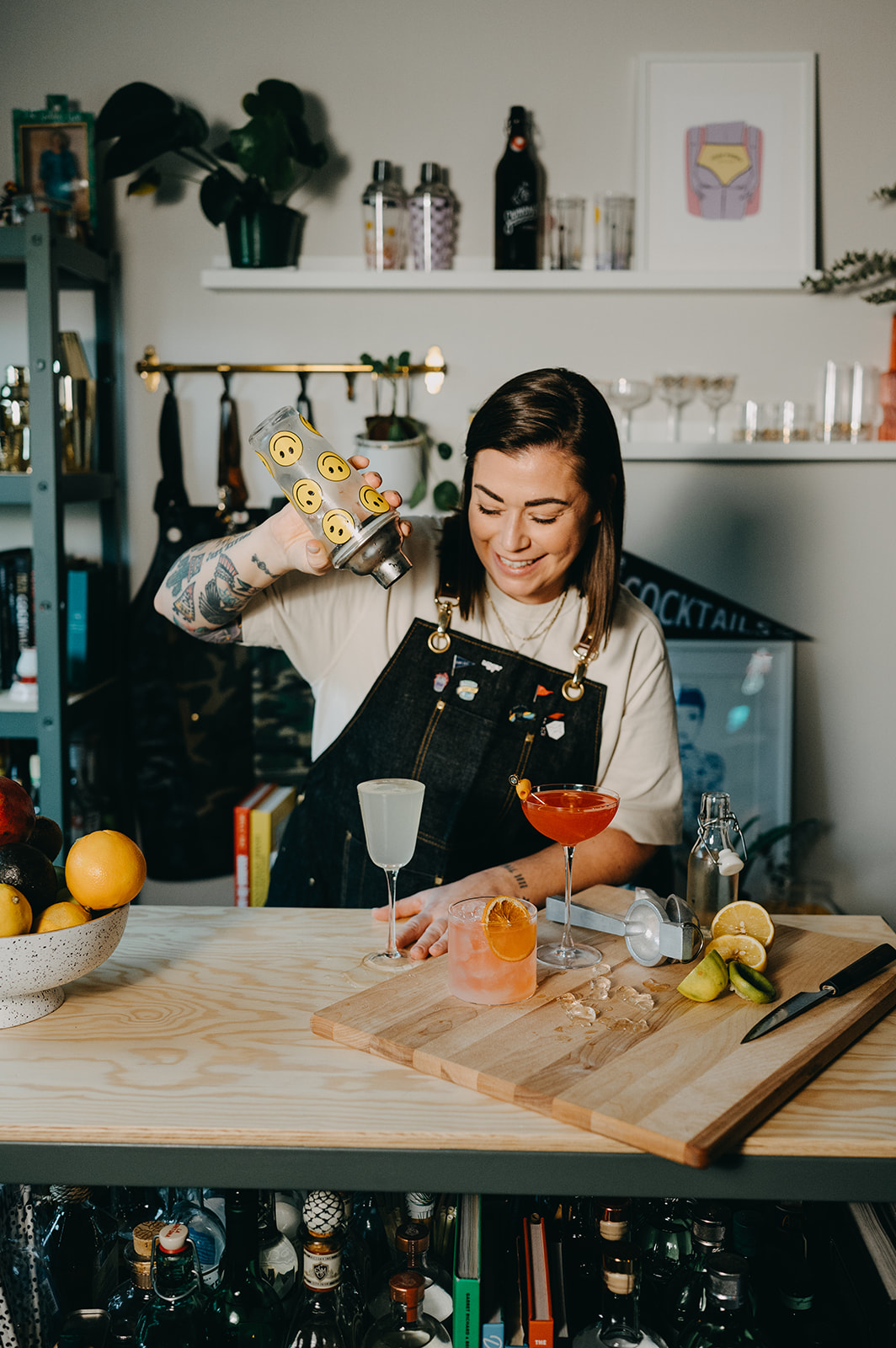 Woman preparing a cocktail behind a bar with bottles, fruits, and bar tools on the counter.
