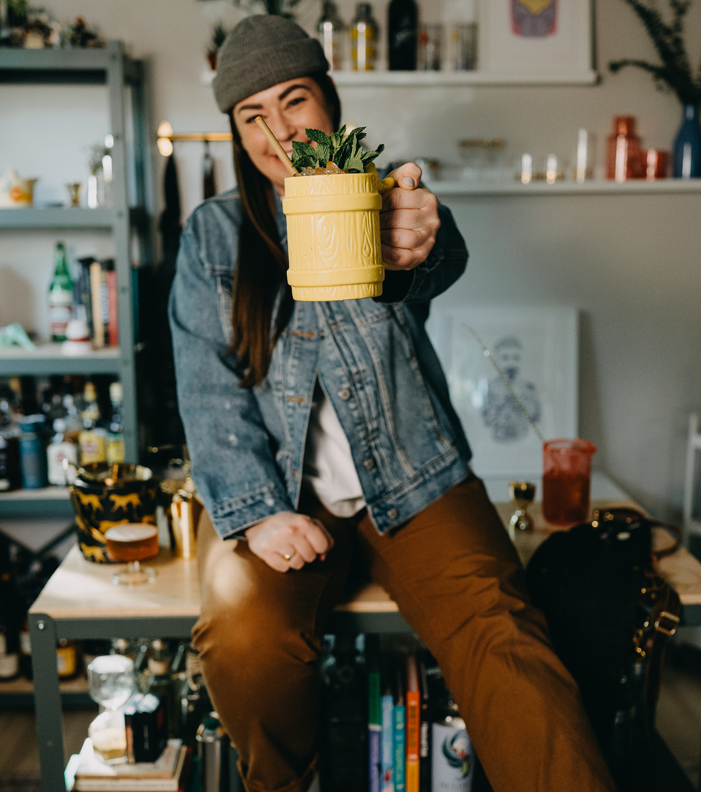 Person holding a yellow mug with a plant, smiling, in a room with shelves and various items, casual clothing.