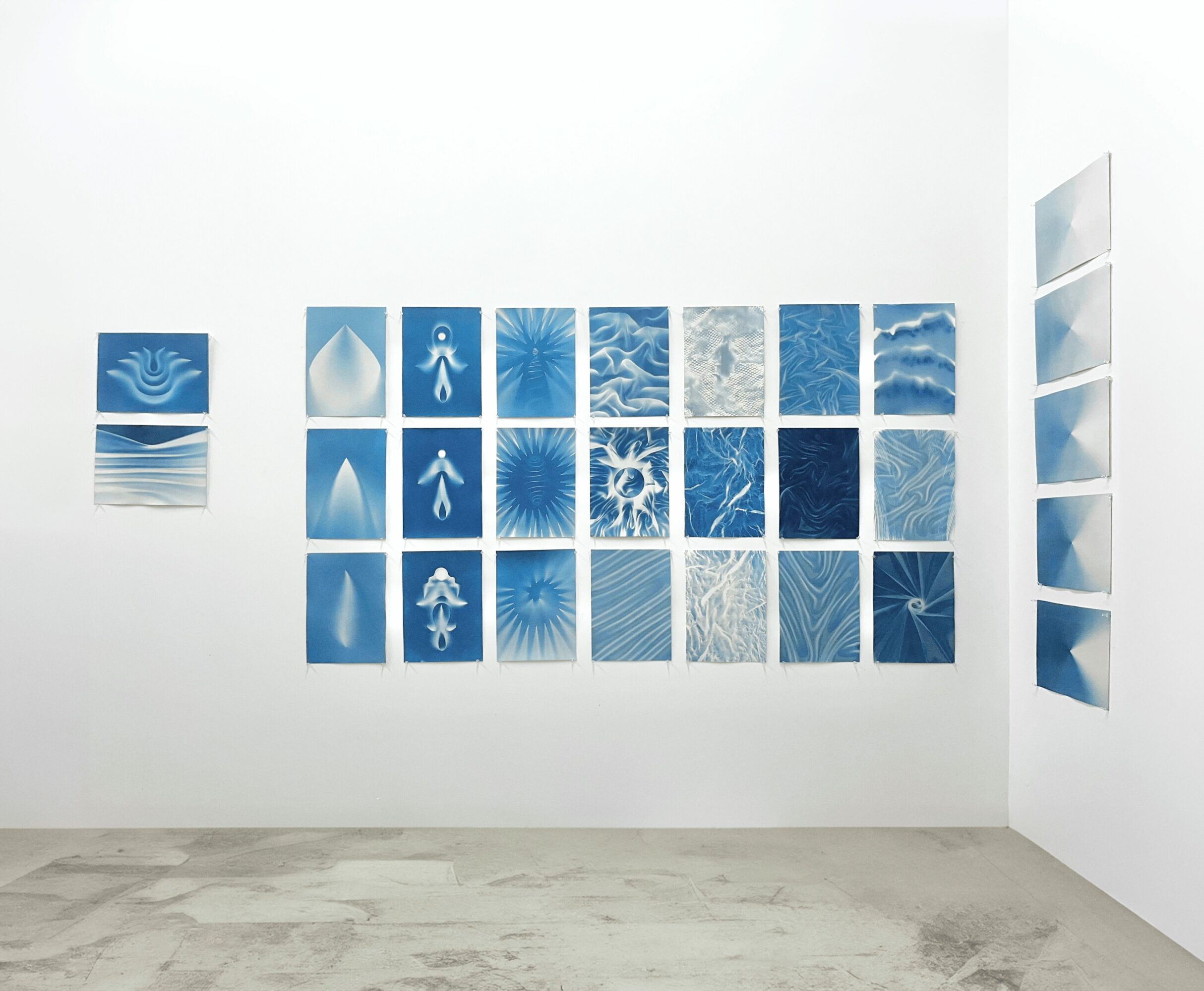 Gallery wall with blue abstract art pieces arranged in a grid, with additional pieces on the right wall, in a white room.