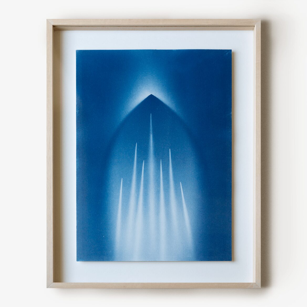 Framed abstract blue artwork with a pyramid shape and light streaks, on a white wall.