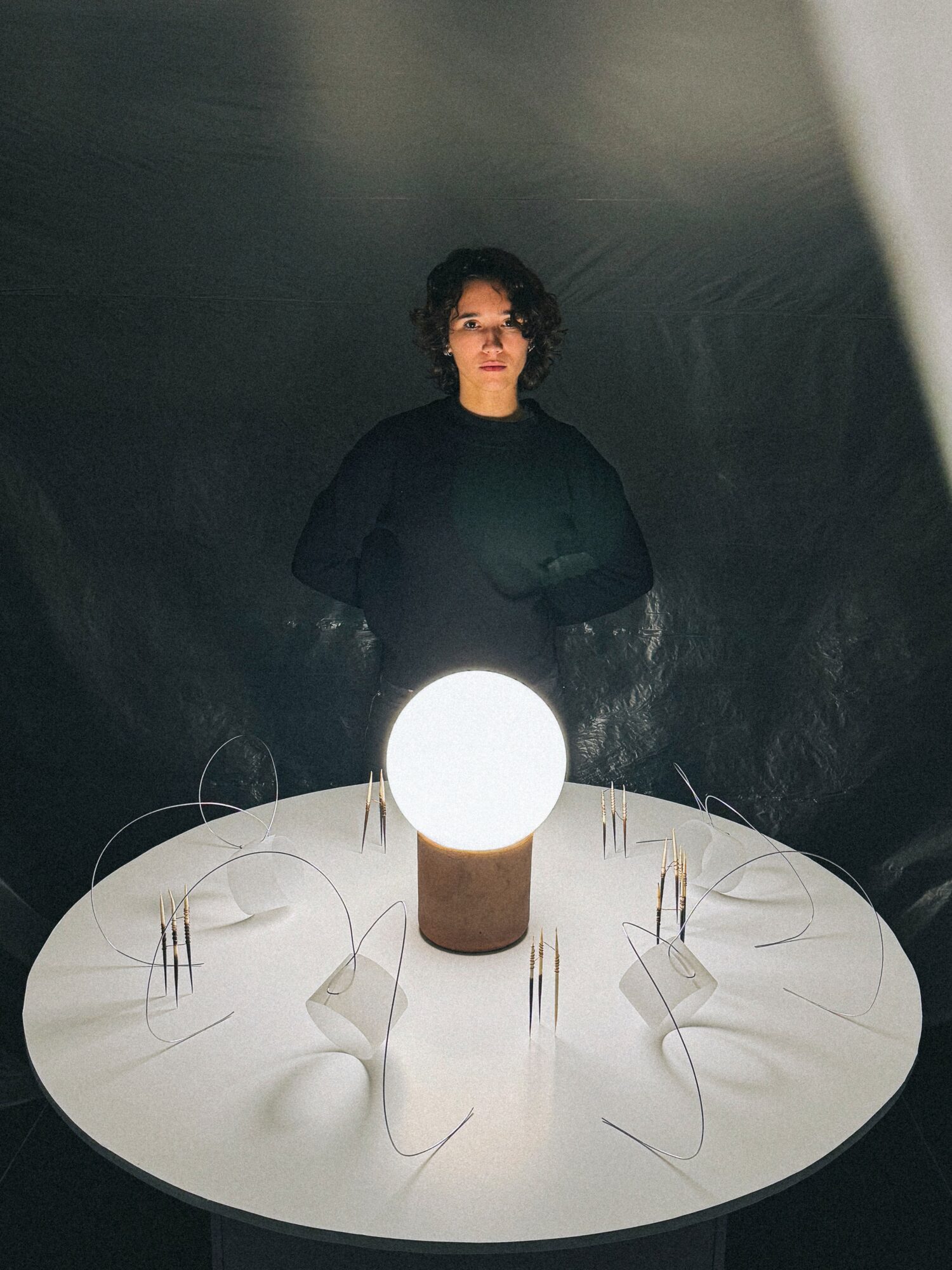 Person with curly hair and glasses stands behind a round table with a glowing orb in the center.