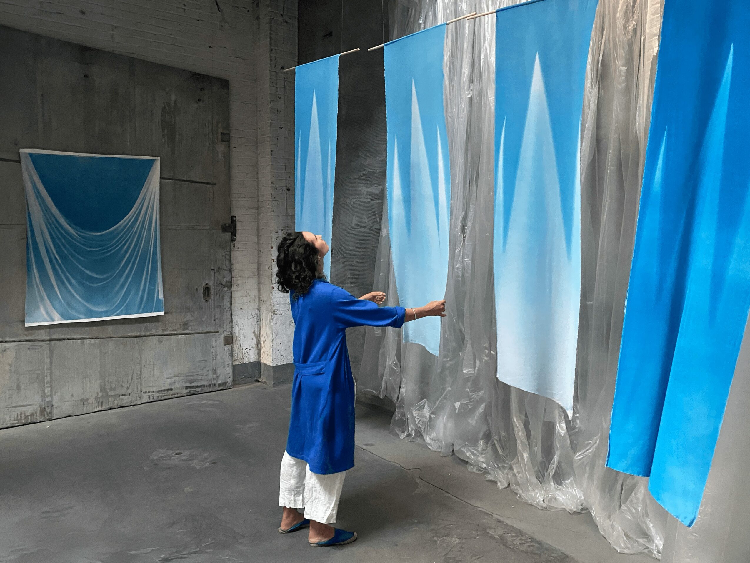 Person in blue coat and white pants interacts with large blue and white fabric panels hanging on wall.