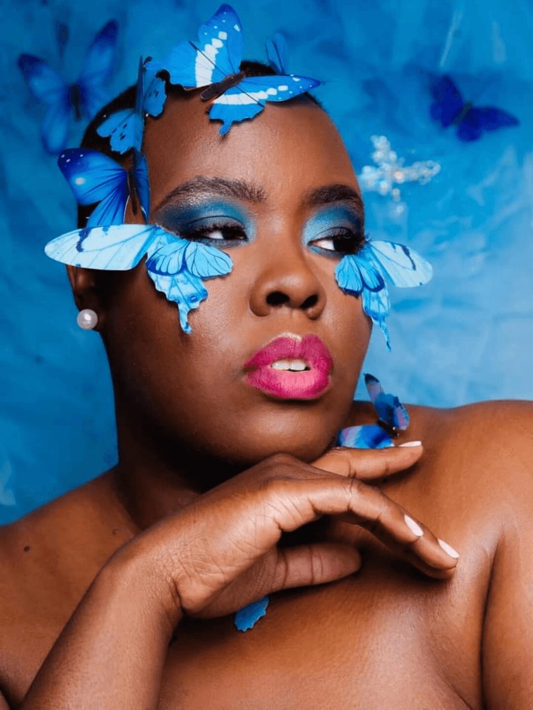 Close-up of a woman with blue butterfly makeup and butterflies on her face, against a blue background.