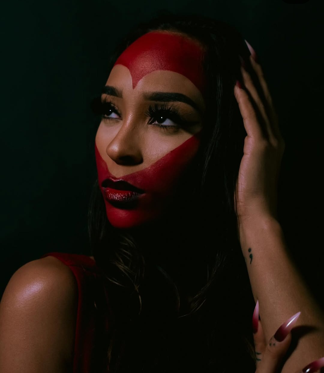 Woman with red face paint, dark hair, and makeup, touching her hair with her hand, dark background