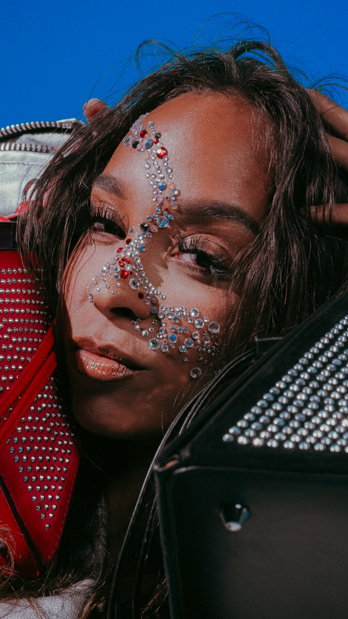 Close-up of a woman with decorative rhinestones on her face, dark hair, and a red and black outfit, against a blue background.