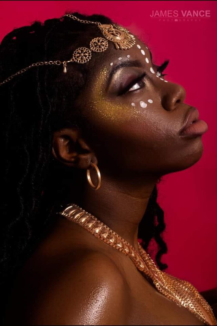 Portrait of a woman with elaborate jewelry and face paint against a pink background.