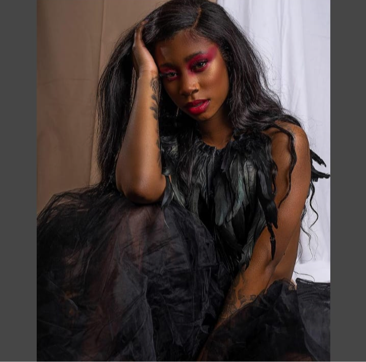 Young woman with long black hair and red makeup, wearing a black feathered top, sitting with her hand on her head.