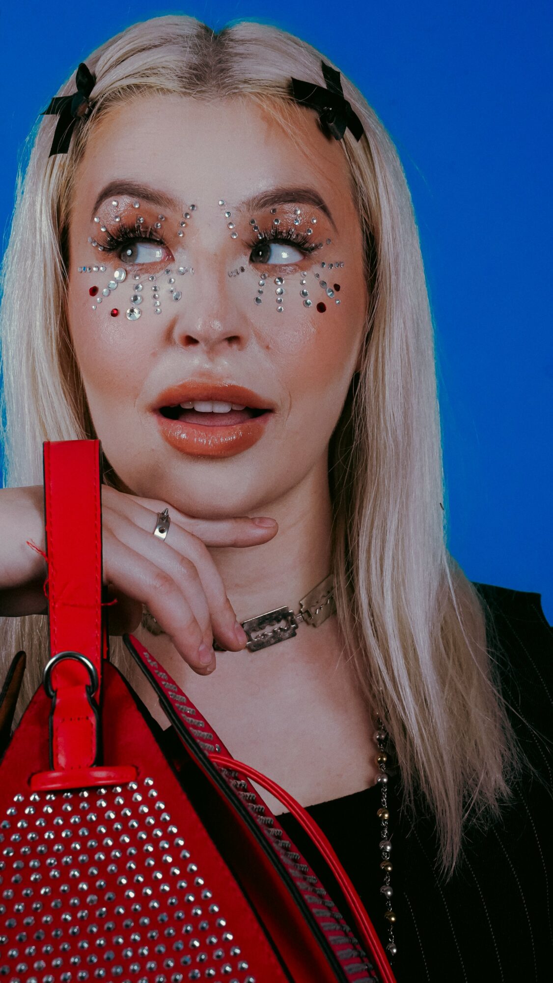 Young woman with long blonde hair, makeup, and decorative face gems, holding a red purse against a blue background.