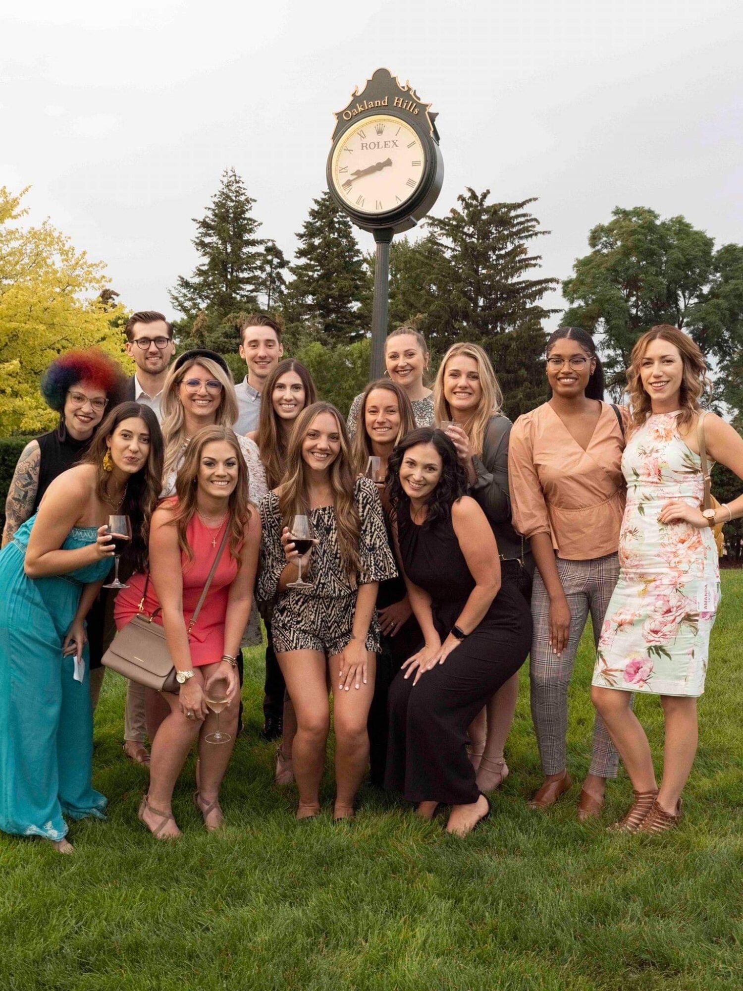 Group of people posing outdoors near a clock, standing on grass with trees in the background.