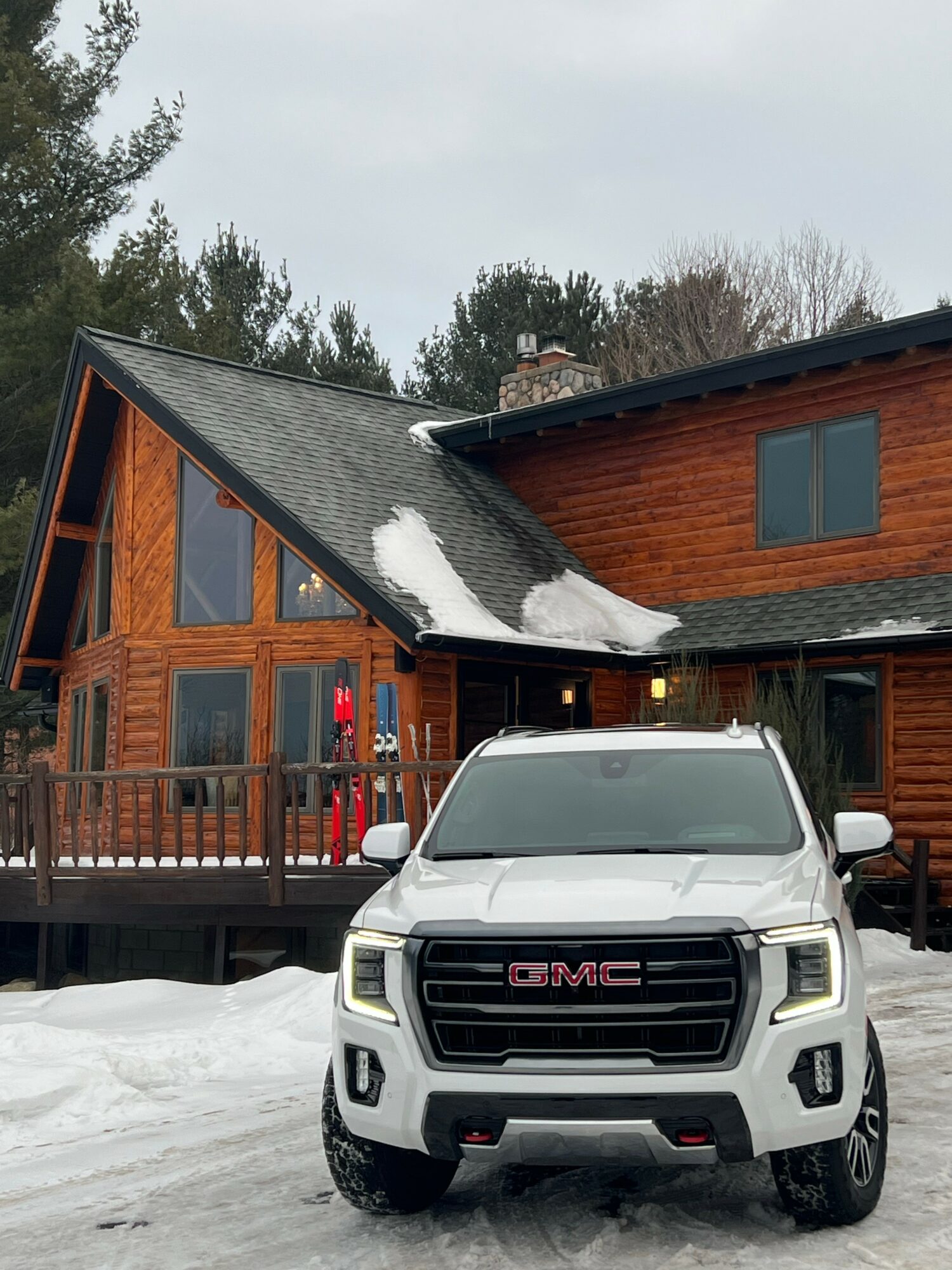 White GMC truck parked in front of a wooden house with snow on the roof and ground.